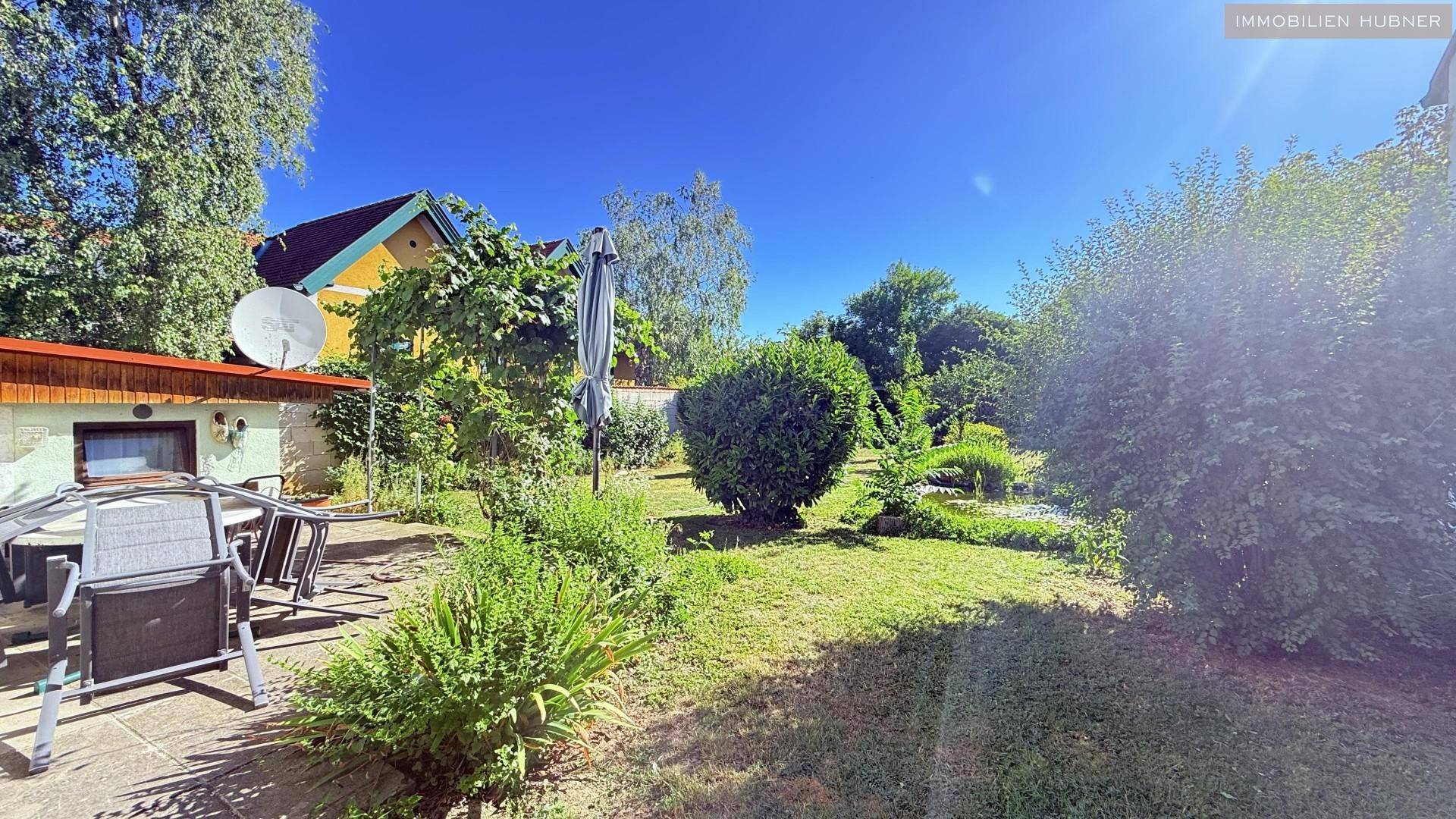 Gepflasterte Terrasse mit Gartenmöbeln und Sonnenschirm, umgeben von Grünflächen.