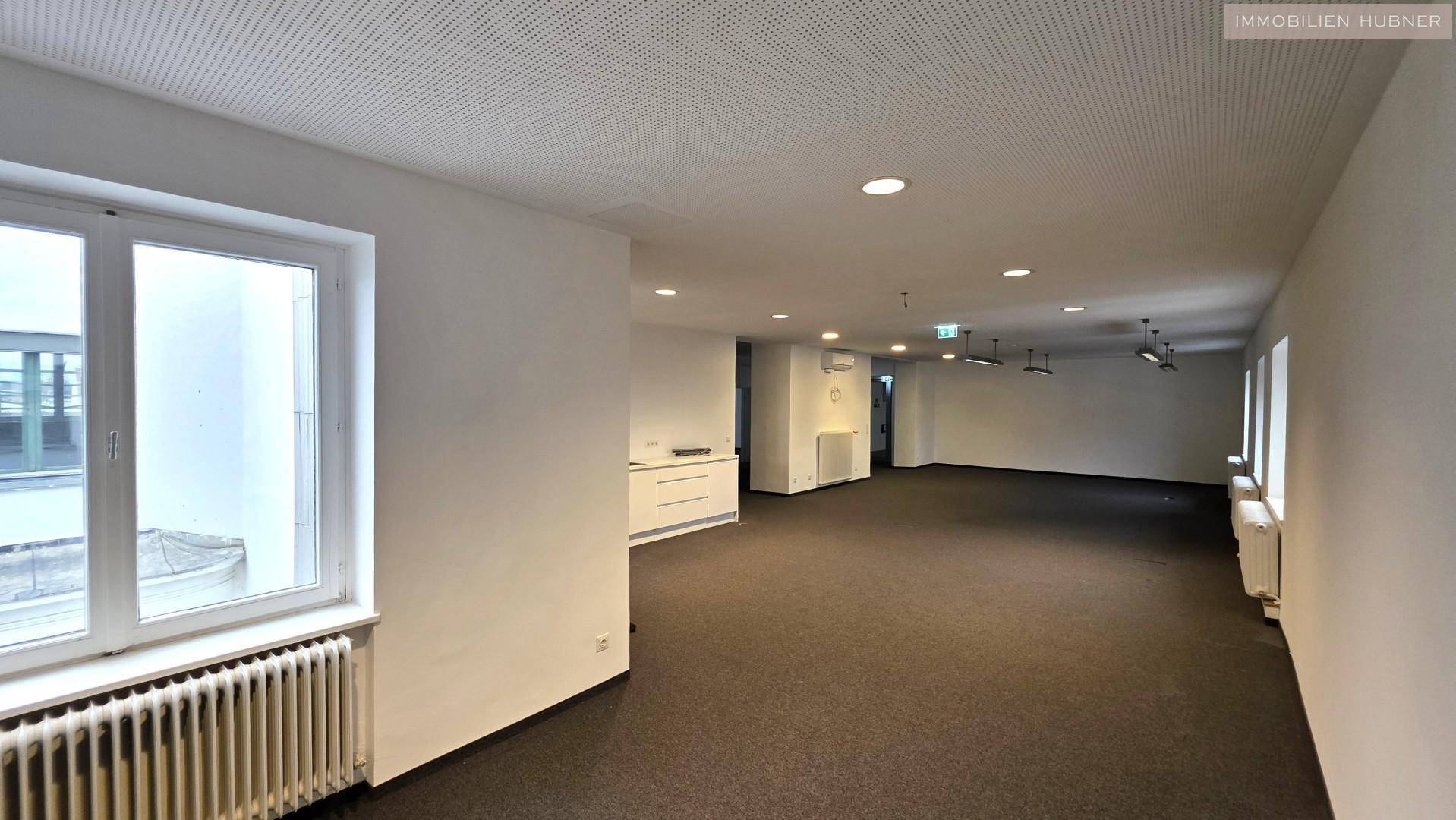 Weiterer Blick in den großen Büroraum mit Teppichboden, Einbauschrank und Fenstern.