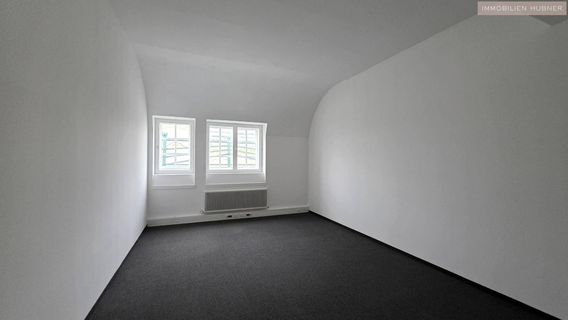Leerer Raum mit schrägen Wänden, dunklem Teppichboden und zwei Fenstern, möglicherweise im Dachgeschoss.