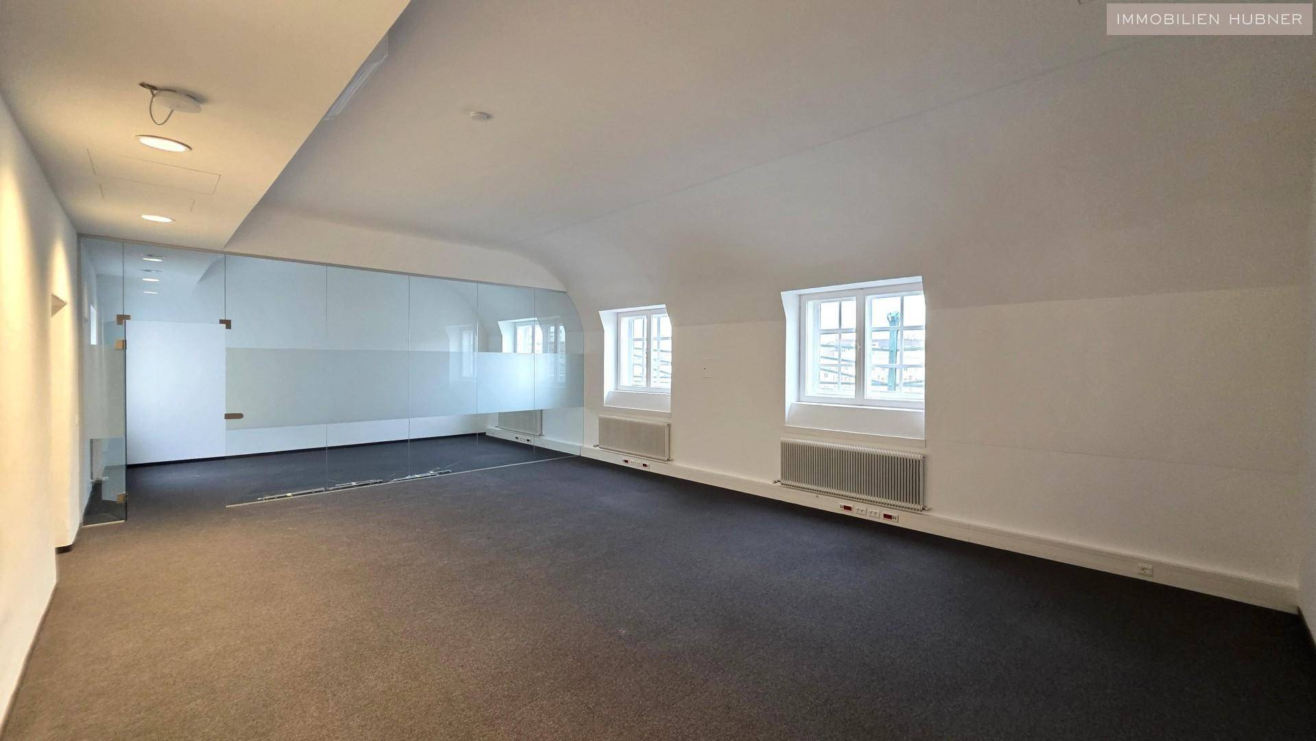 Helles Büro mit Dachschrägen, Teppichboden und einer modernen Glasabtrennung.