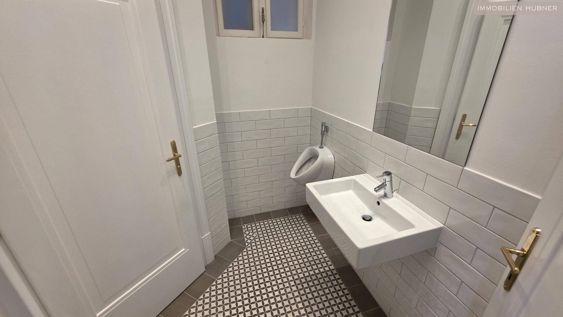 Modernes Gäste-WC mit Urinal, Waschbecken und einem kleinen Fenster, hell gefliest.