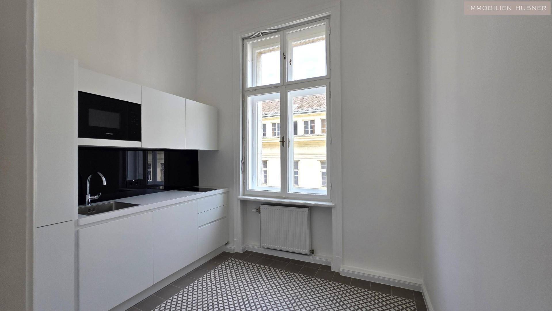 Helle, funktionale Küche mit weißen Fronten, schwarzer Rückwand und einem großen Fenster für Tageslicht.