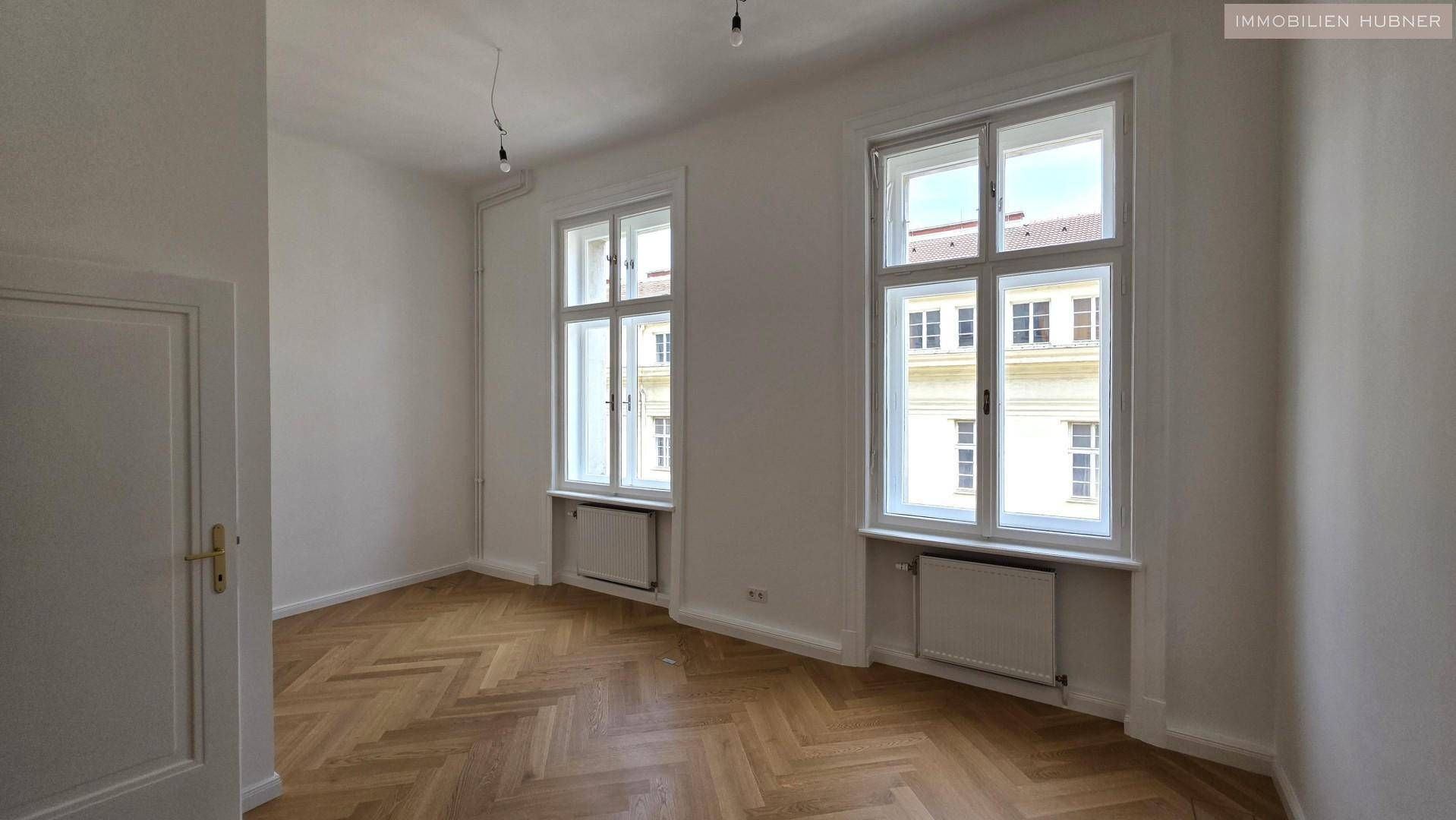 Leerer Raum mit zwei großen Fenstern, die viel natürliches Licht spenden, und elegantem Fischgrätparkett.
