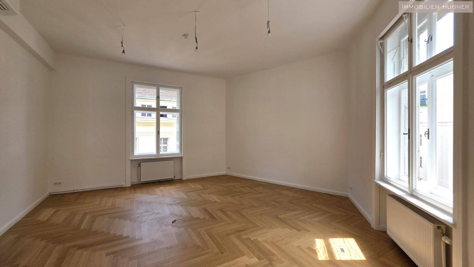 Heller, leerer Raum mit klassischem Fischgrätparkett und großen Fenstern, die viel Tageslicht hereinlassen.