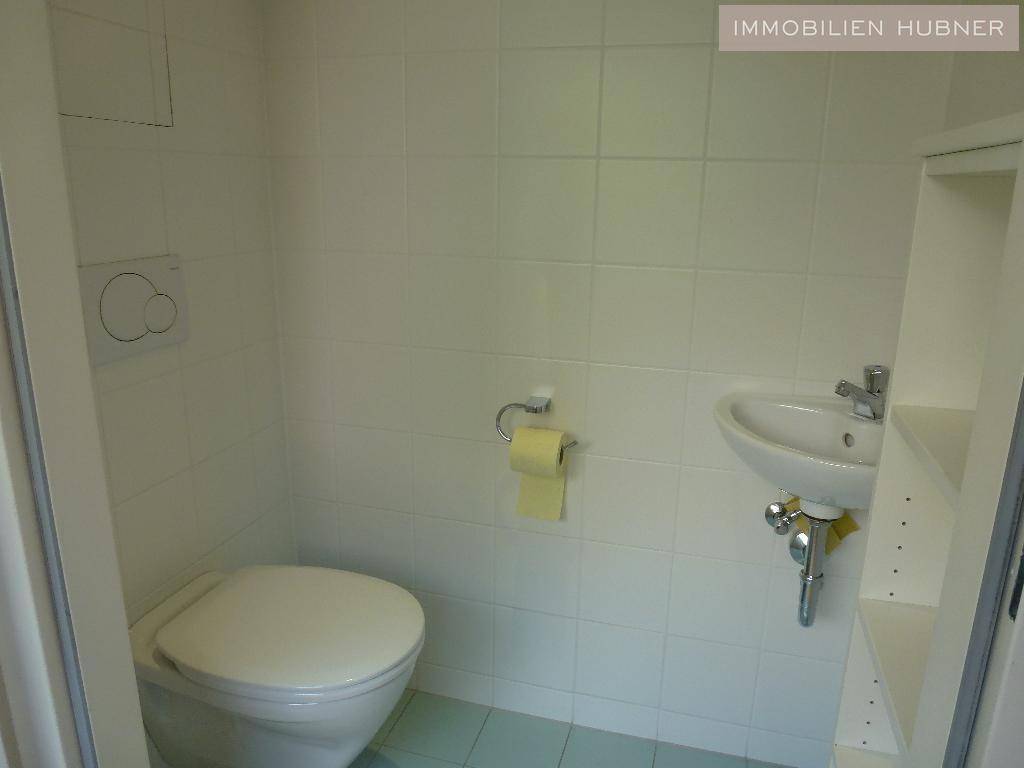 Separates WC mit weißen Fliesen, wandhängender Toilette und einem kleinen Handwaschbecken.
