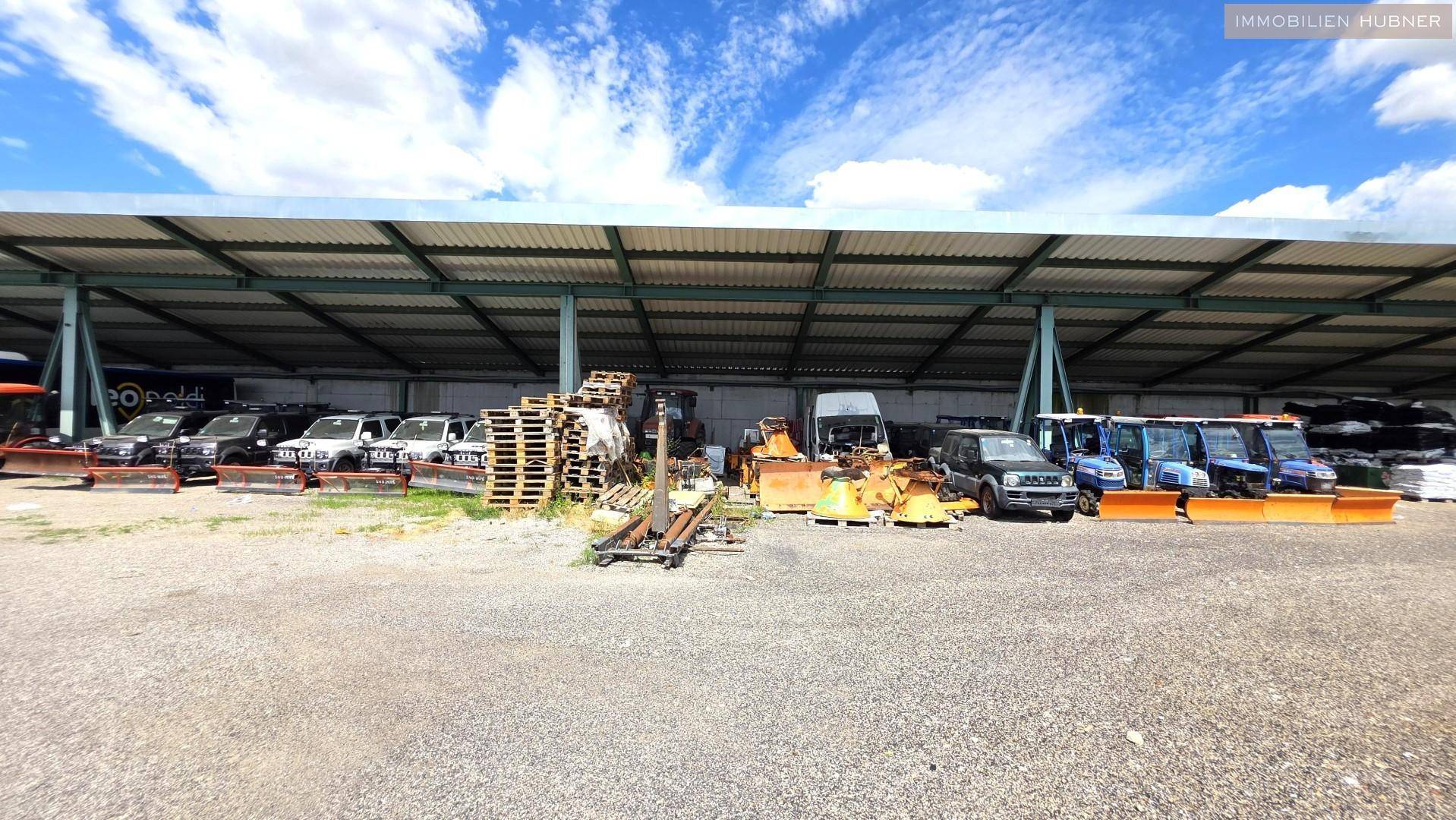 Überdachter Außenbereich mit geparkten Fahrzeugen und gelagerten Paletten unter blauem Himmel.