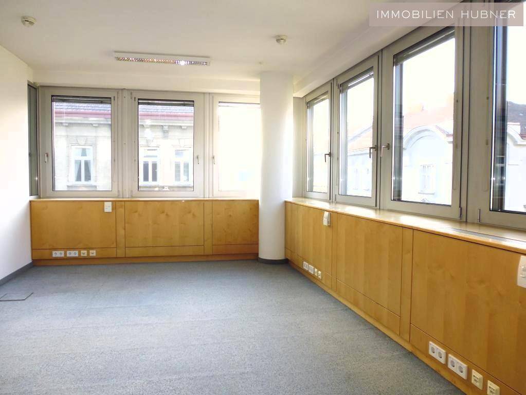 Helles Büro mit großen Fenstern, Teppichboden und integrierten Holzschränken entlang der Wand.