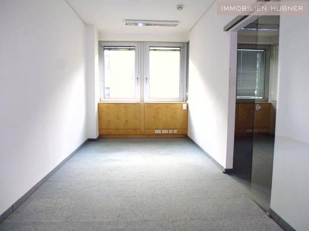 Kleines, helles Büro mit zwei Fenstern, Teppichboden und Holzschränken, daneben eine Glaswand.