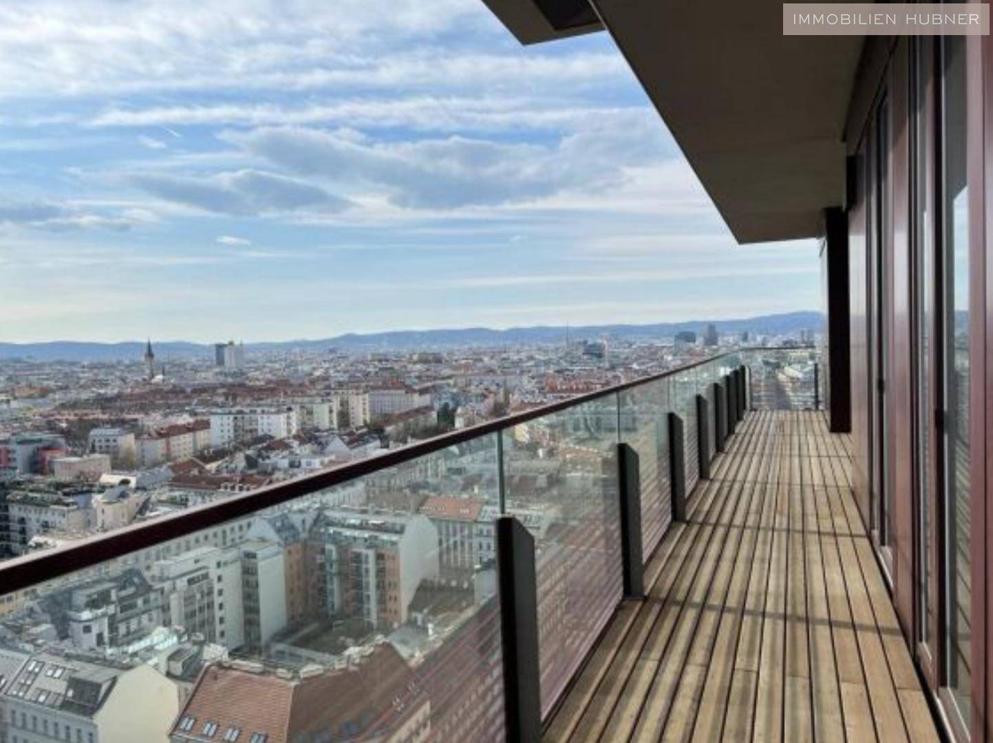Langer Balkon mit Holzbelag und Glasgeländer, der einen beeindruckenden Panoramablick über die Stadt bietet.