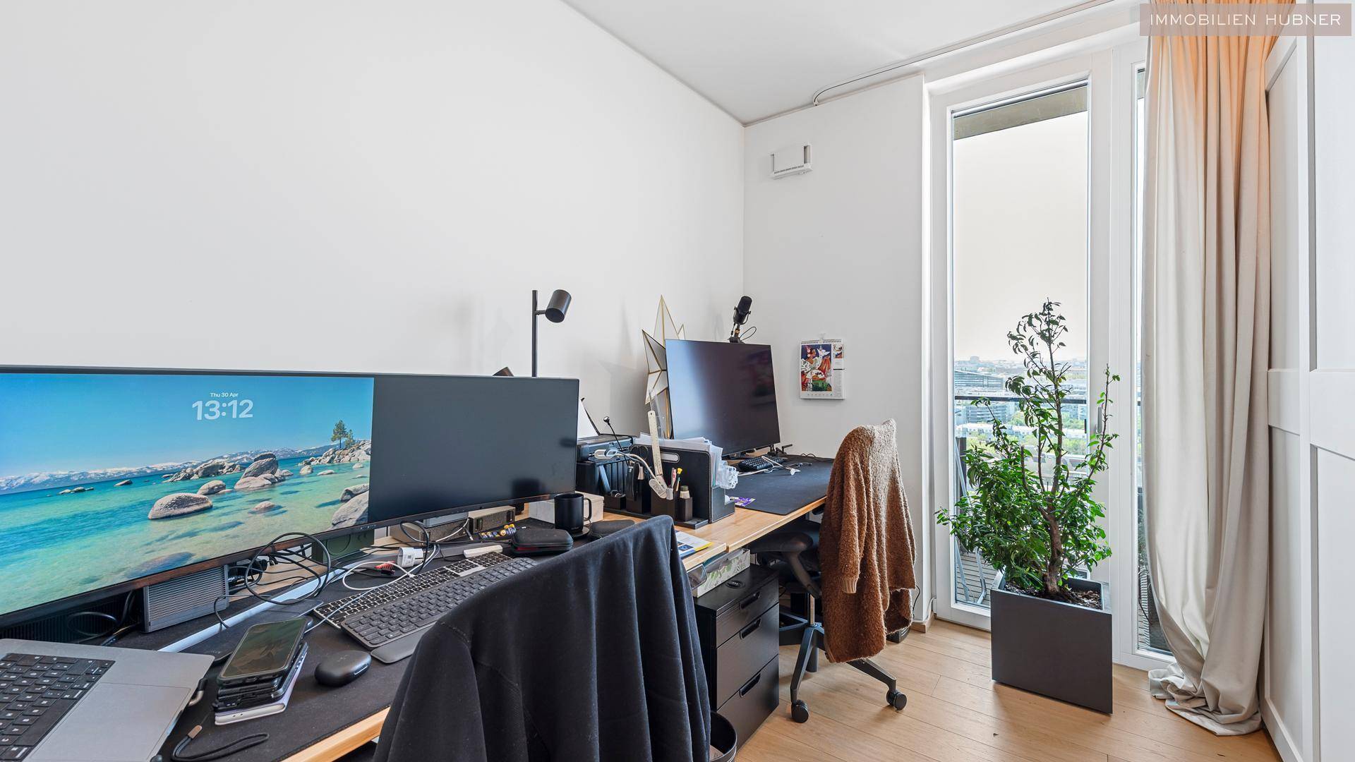 Modernes Heimbüro mit mehreren Bildschirmen und einem Fenster, das Tageslicht hereinlässt.
