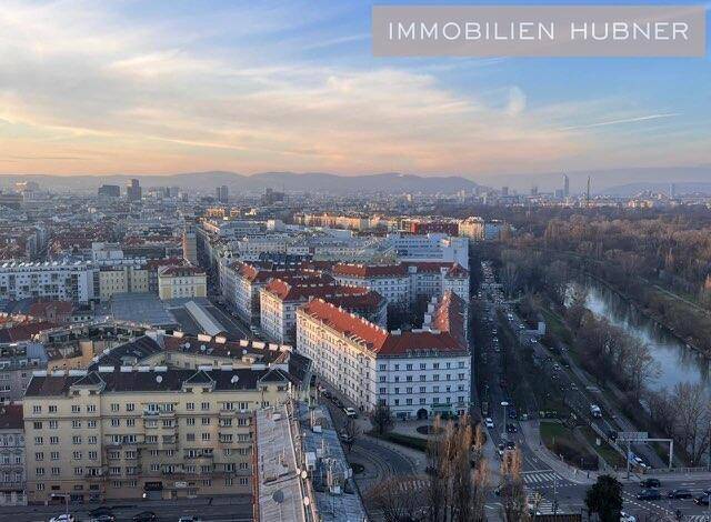 Weitläufiger Blick über die Stadt mit Flusslauf und Bergen am Horizont unter einem bewölkten Himmel.