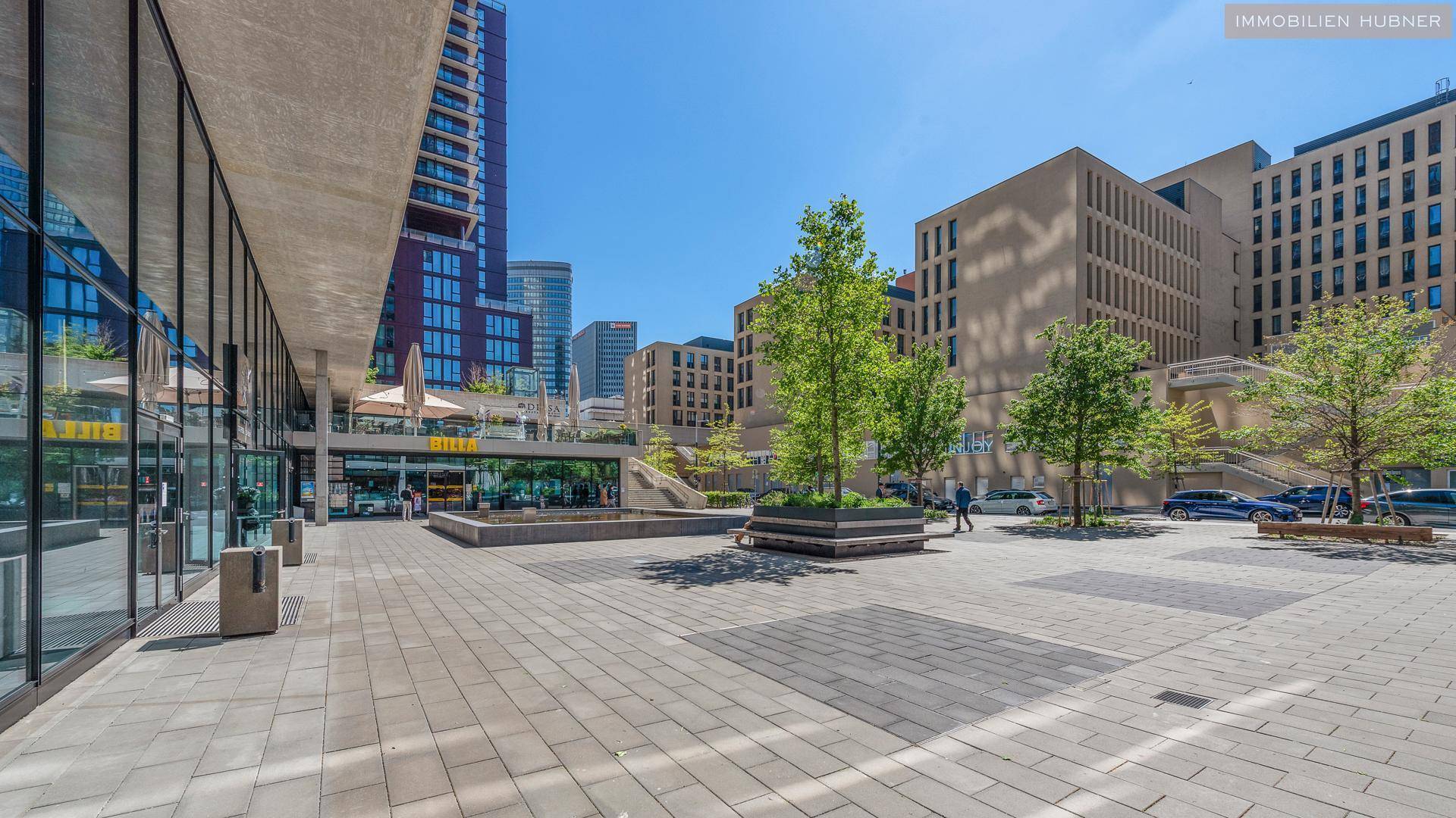 Moderner Stadtplatz mit zeitgenössischer Architektur, großen Glasfassaden und grünen Bäumen unter blauem Himmel.