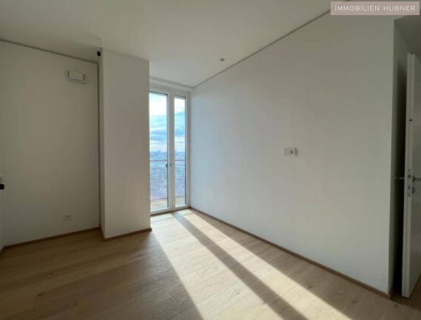 Heller, leerer Raum mit Holzboden und großem Fenster, das viel Tageslicht hereinlässt.