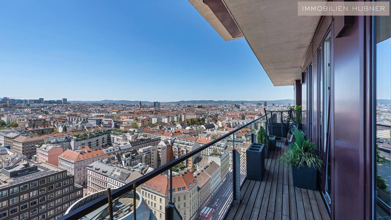 Geräumiger Balkon mit Holzbelag und Glasgeländer, der einen weiten Blick über die Stadt ermöglicht.