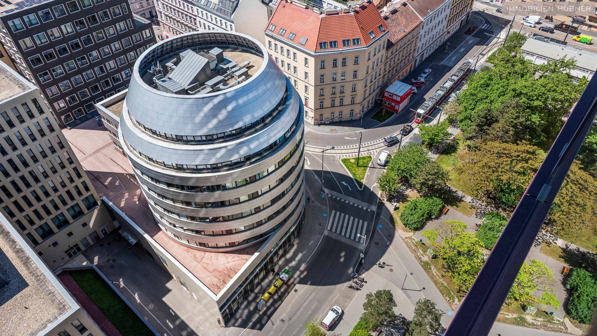 Luftaufnahme eines modernen zylindrischen Gebäudes inmitten einer städtischen Umgebung mit Straßen und Grünflächen.