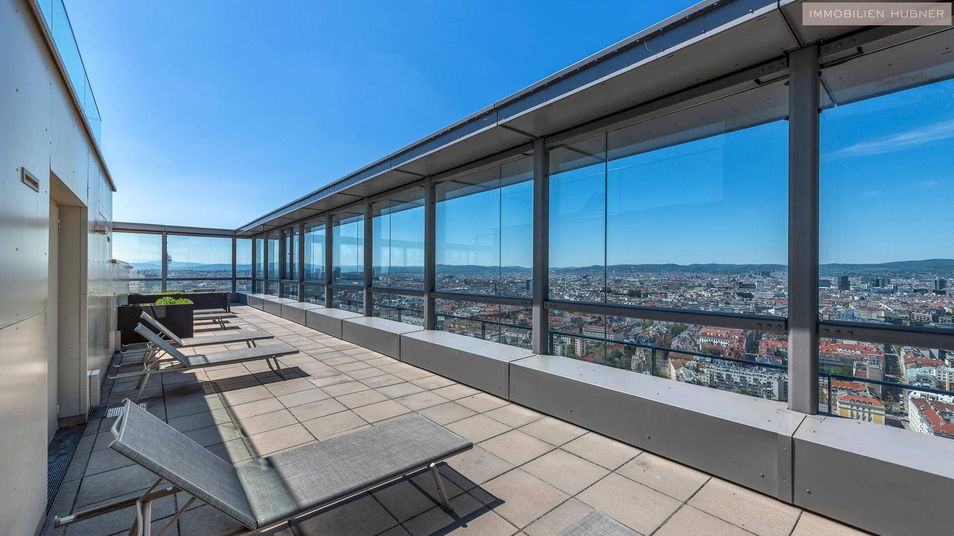 Große Dachterrasse mit Liegestühlen und Glaswänden, die einen weiten Blick über die Stadt bietet.