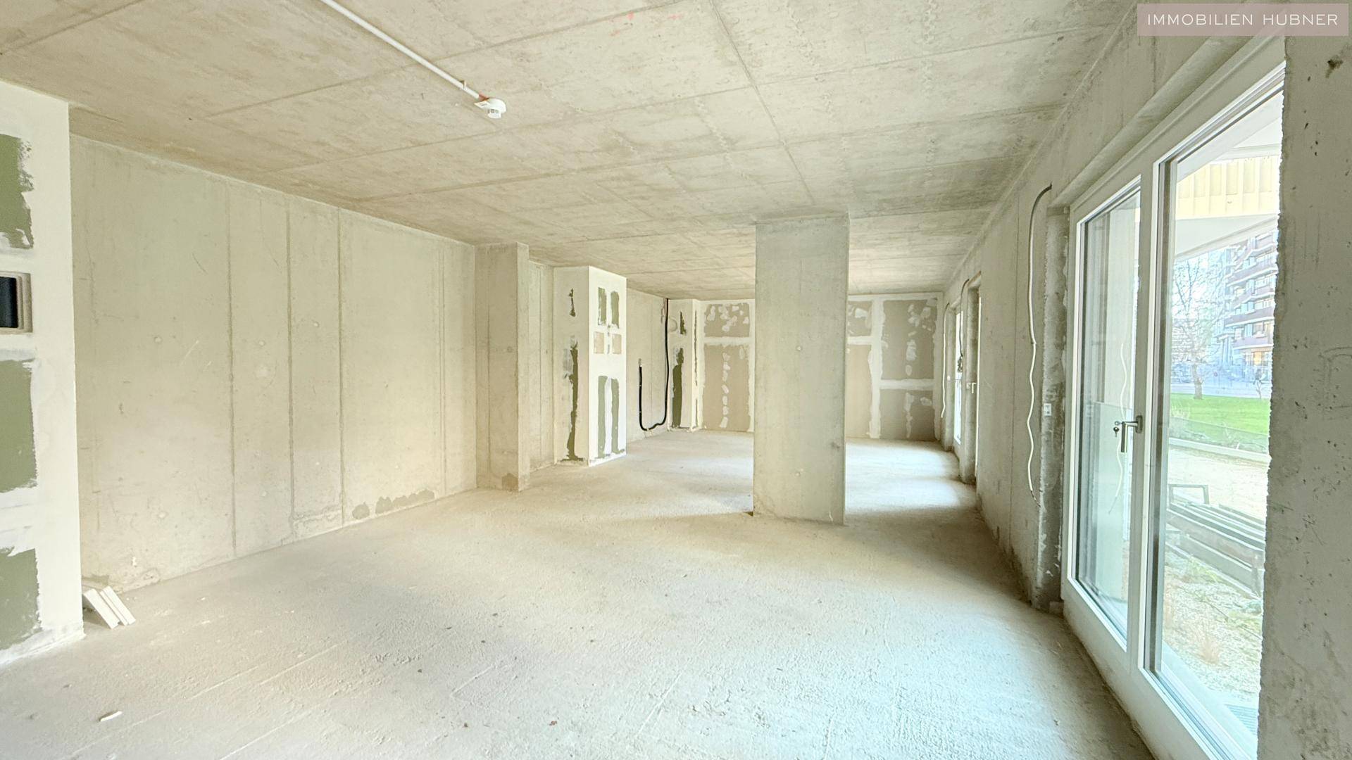 Großer, unfertiger Raum mit Betonboden und vorbereiteten Wänden, viel Tageslicht durch Fenster.