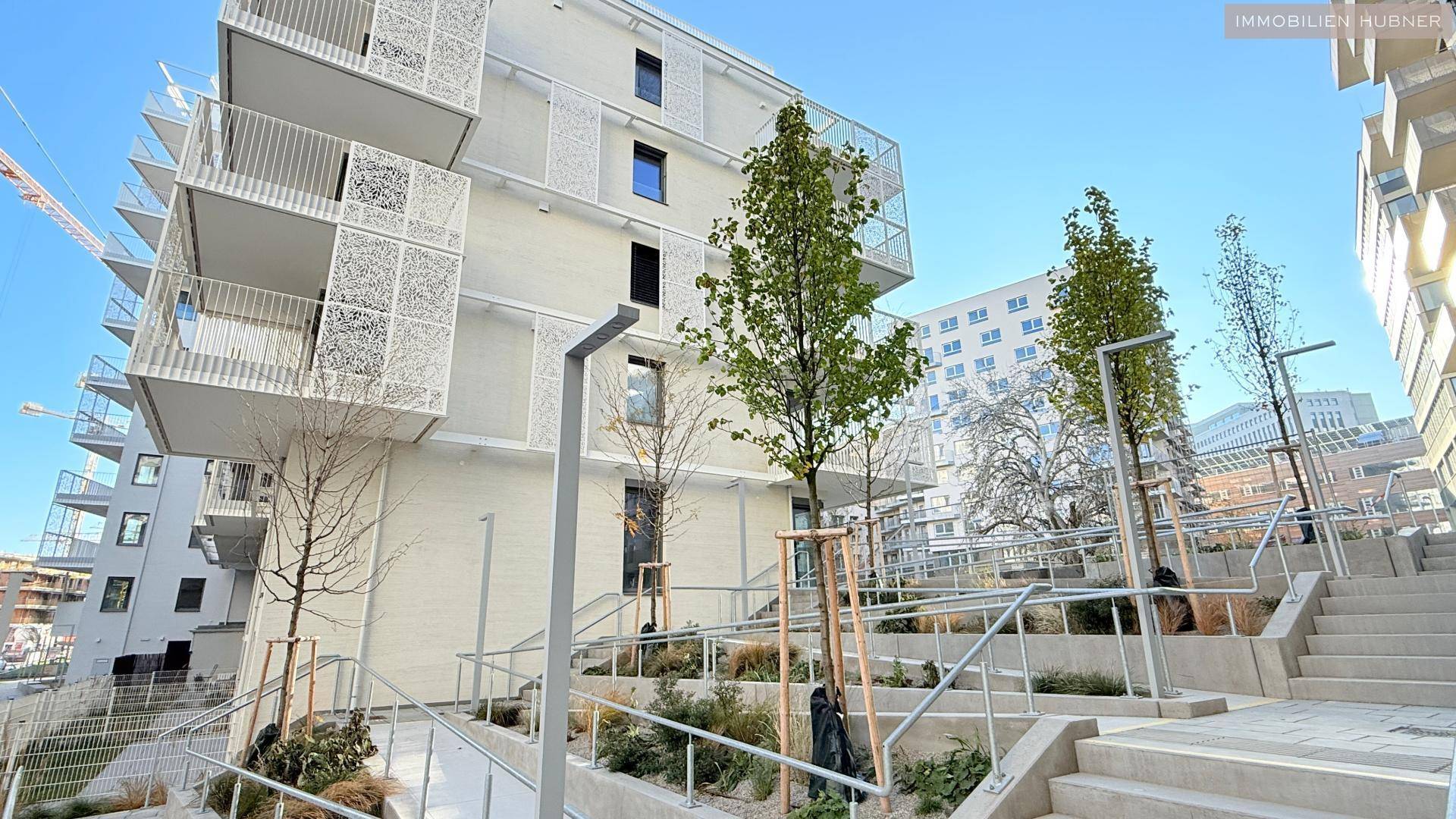 Außenansicht eines modernen Gebäudes mit Terrassen, Treppen und Grünflächen im Eingangsbereich.