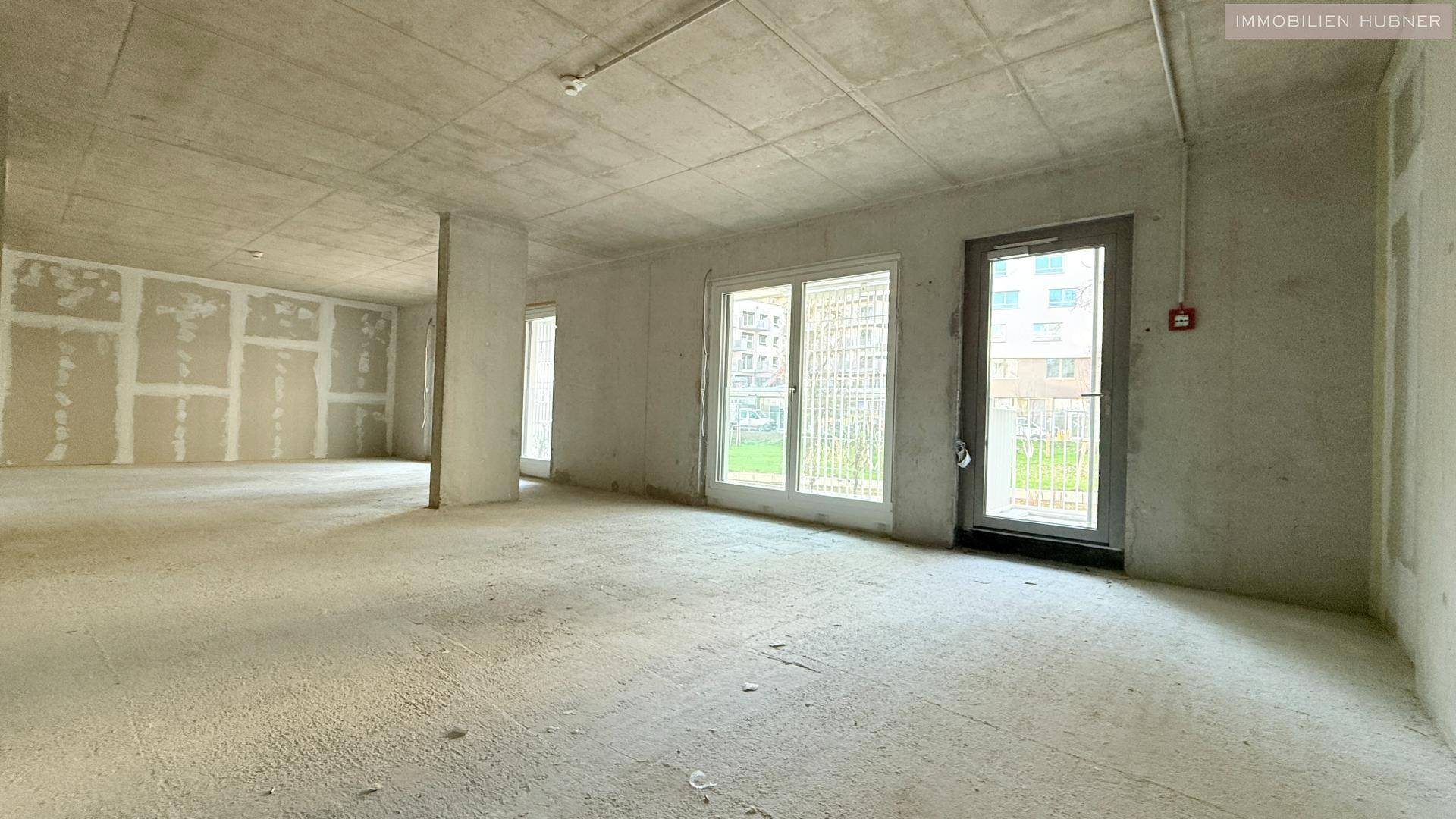 Unfertiger Innenraum mit Betonwänden und großen Fenstern, bereit für den Ausbau.
