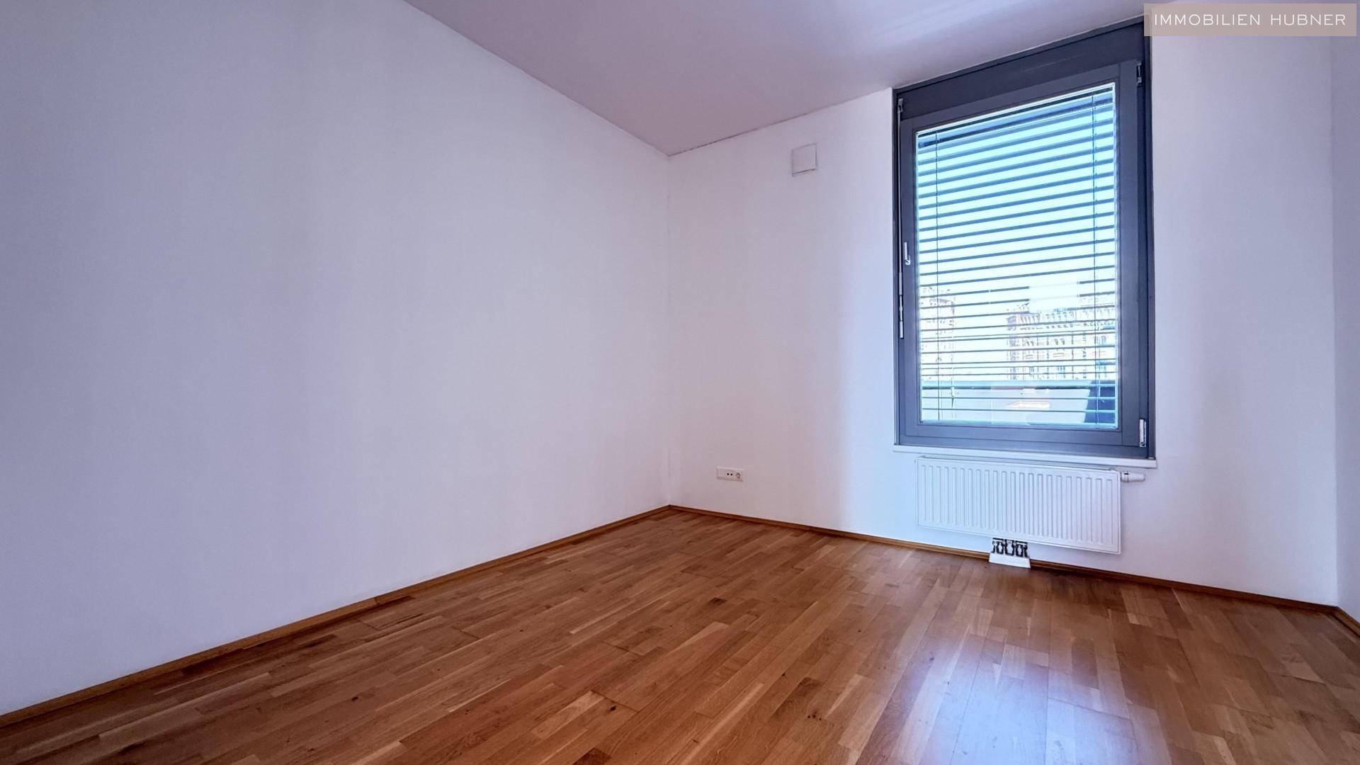Ruhiges Schlafzimmer mit Holzboden, weißen Wänden und einem Fenster, das viel Tageslicht hereinlässt.