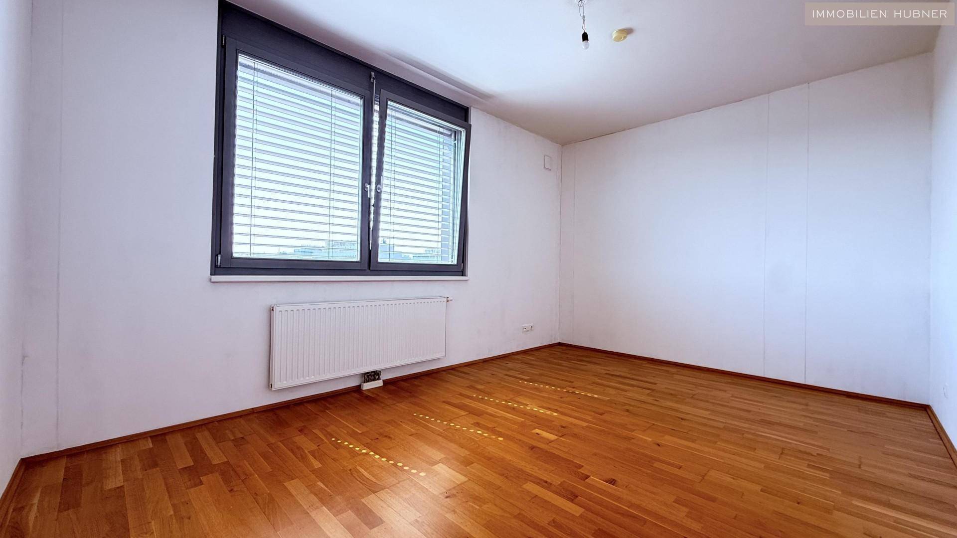 Helles Schlafzimmer mit Parkettboden, weißen Wänden und einem großen Fenster mit Jalousien.