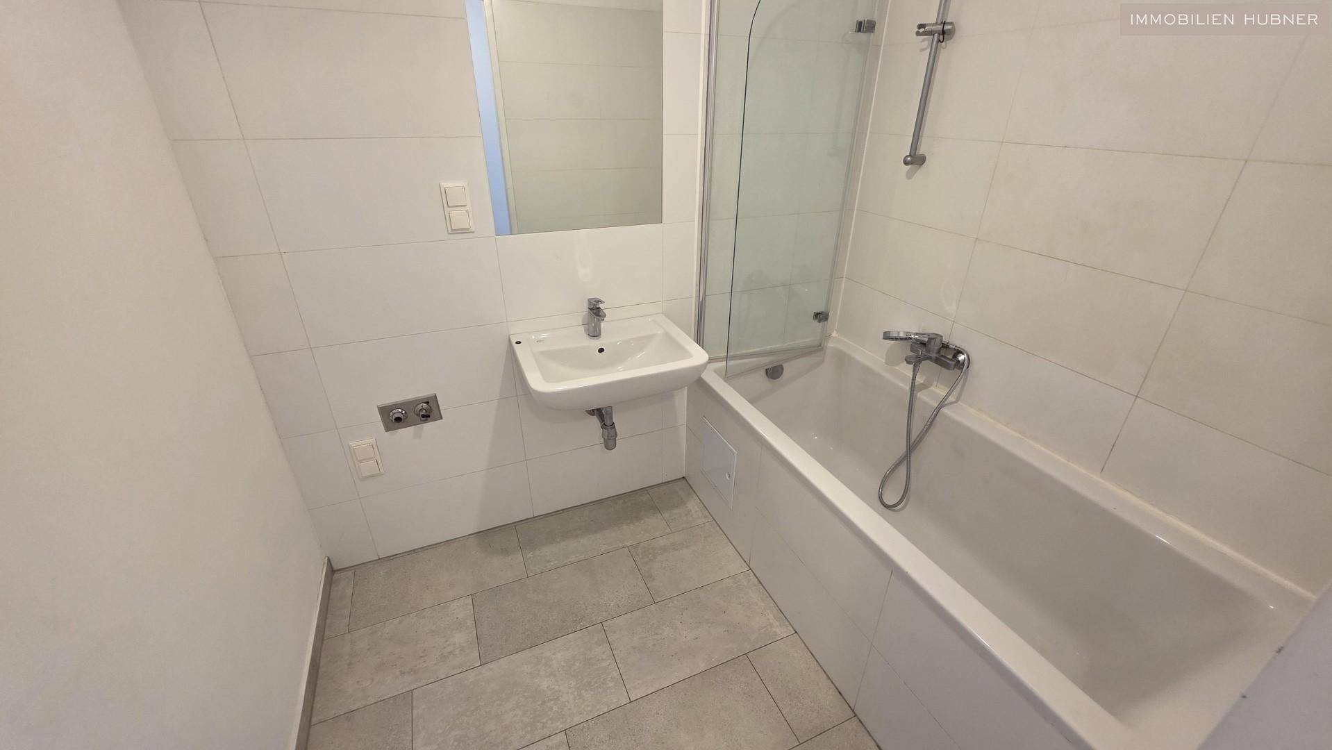 Modernes Badezimmer mit weißen Wandfliesen, grauen Bodenfliesen und Badewanne mit Duschabtrennung.