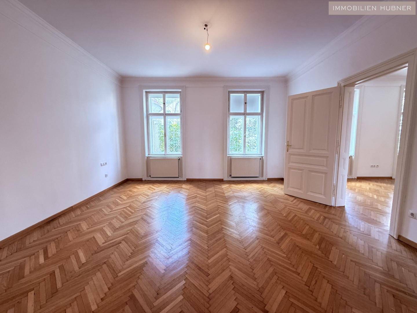 Geräumiges Zimmer mit klassischem Fischgrätparkett und zwei großen Fenstern.