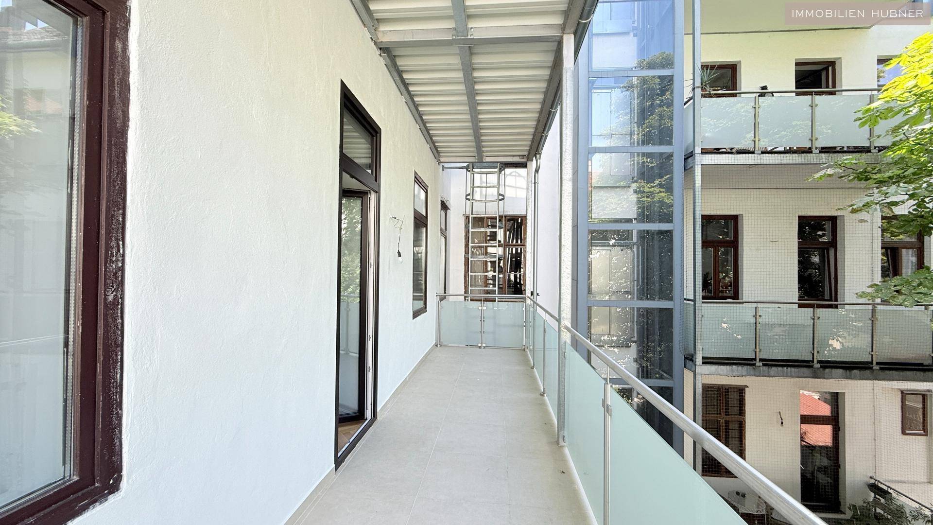 Langer Balkon mit Fliesenboden, Glasgeländer und einem sichtbaren Glasaufzug am Gebäude.
