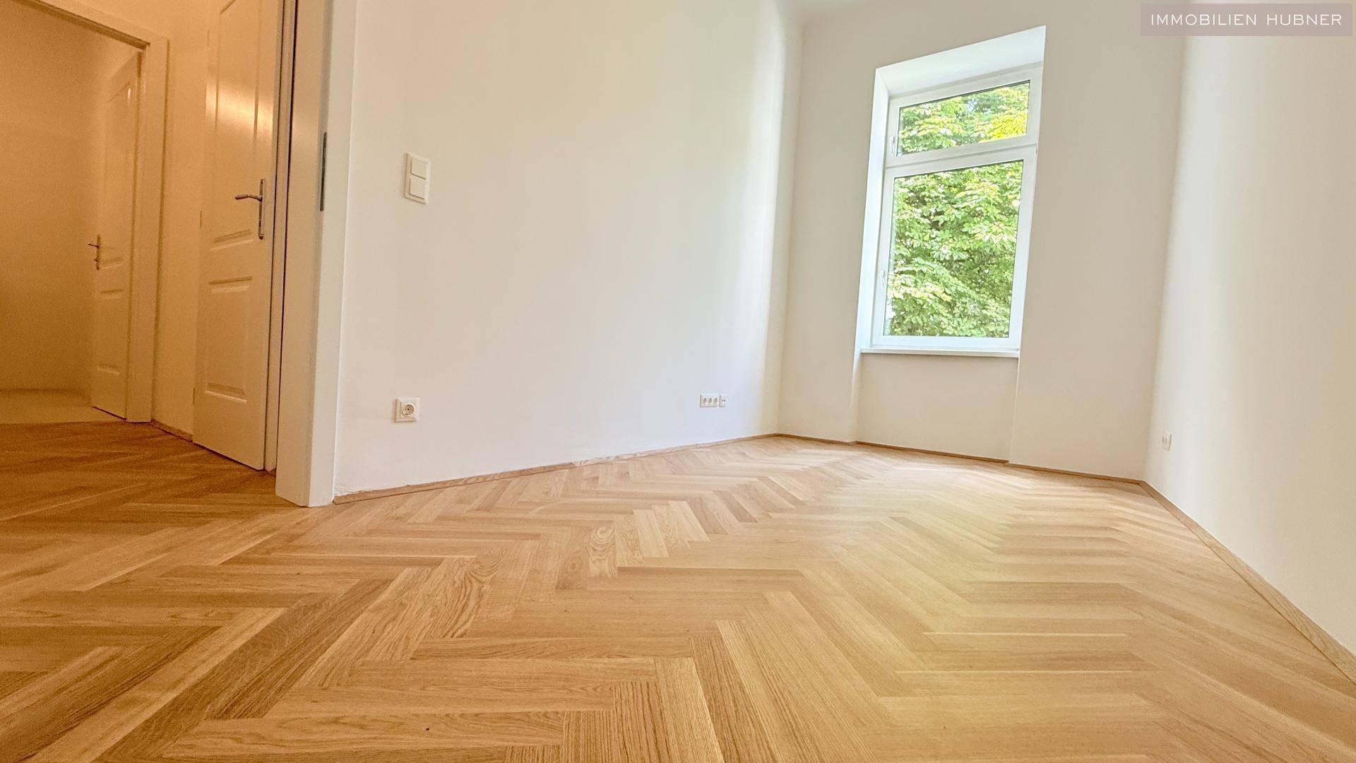 Helles Zimmer mit Fischgrätparkettboden, weißen Wänden und Blick ins Grüne.