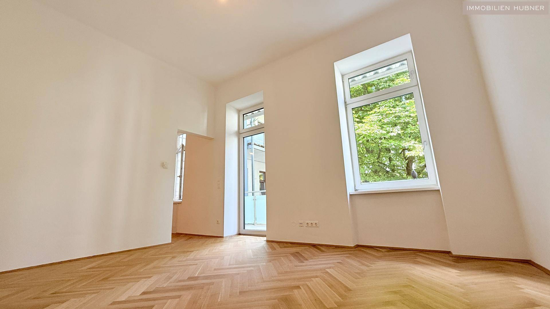 Geräumiger Raum mit Fischgrätparkettboden, großen Fenstern und Zugang zum Balkon.
