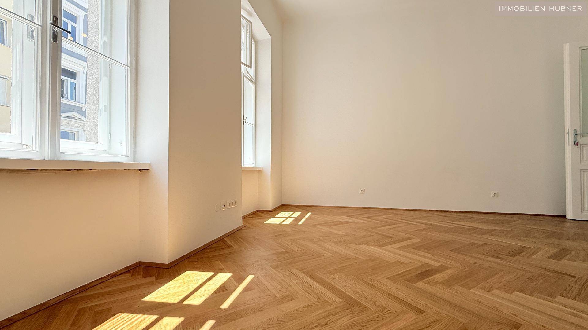 Sonniges Eckzimmer mit Fischgrätparkett und großen Fenstern, die viel natürliches Licht hereinlassen.