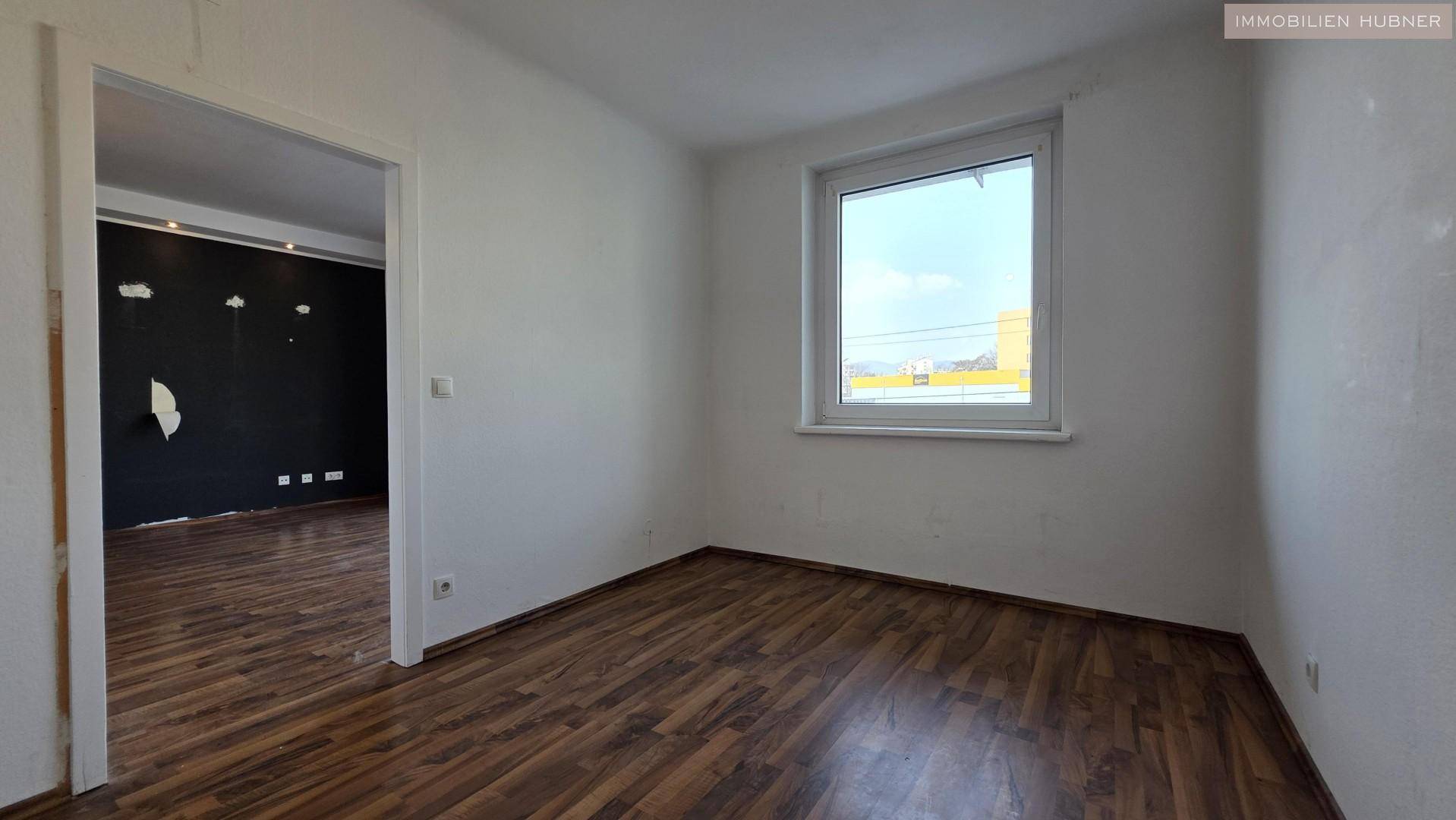 Helles Schlafzimmer mit Holzboden und Fenster, angrenzend an einen weiteren Wohnbereich.