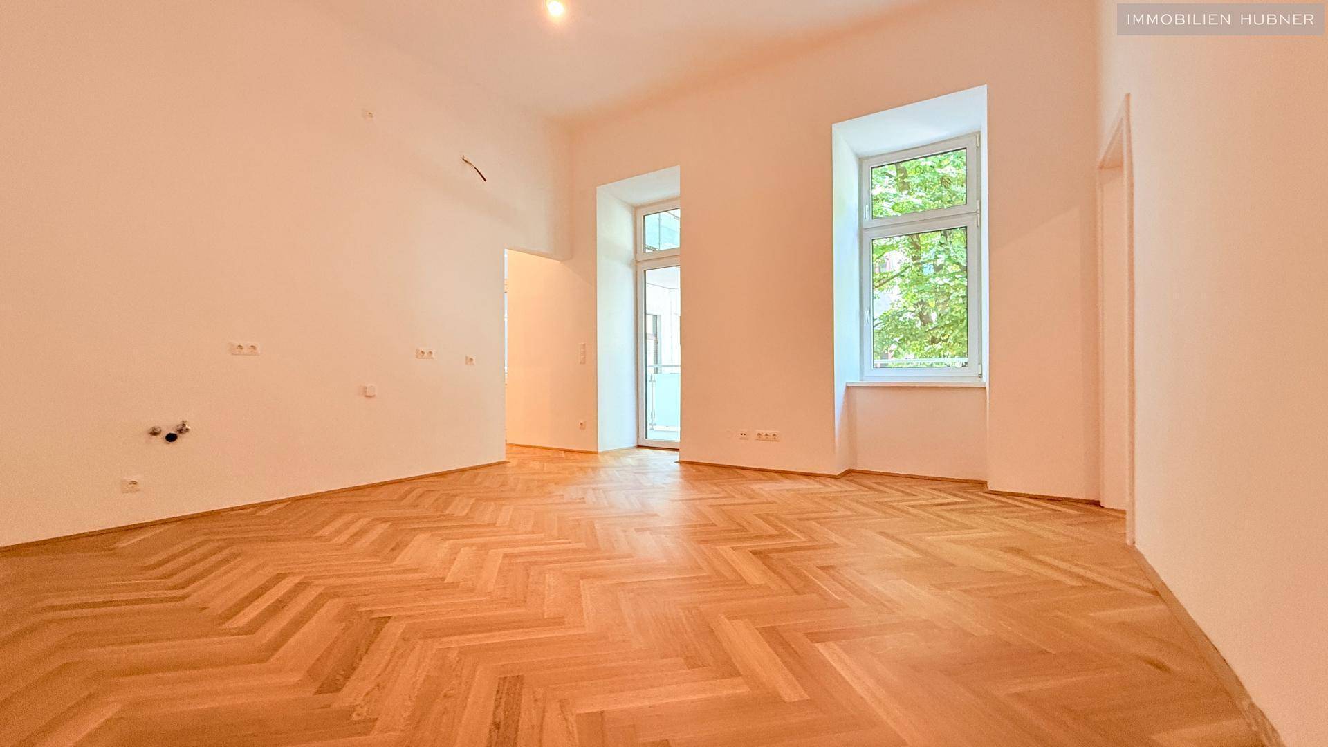 Helles, leeres Zimmer mit Fischgrätparkettboden, hohen Decken und großen Fenstern.