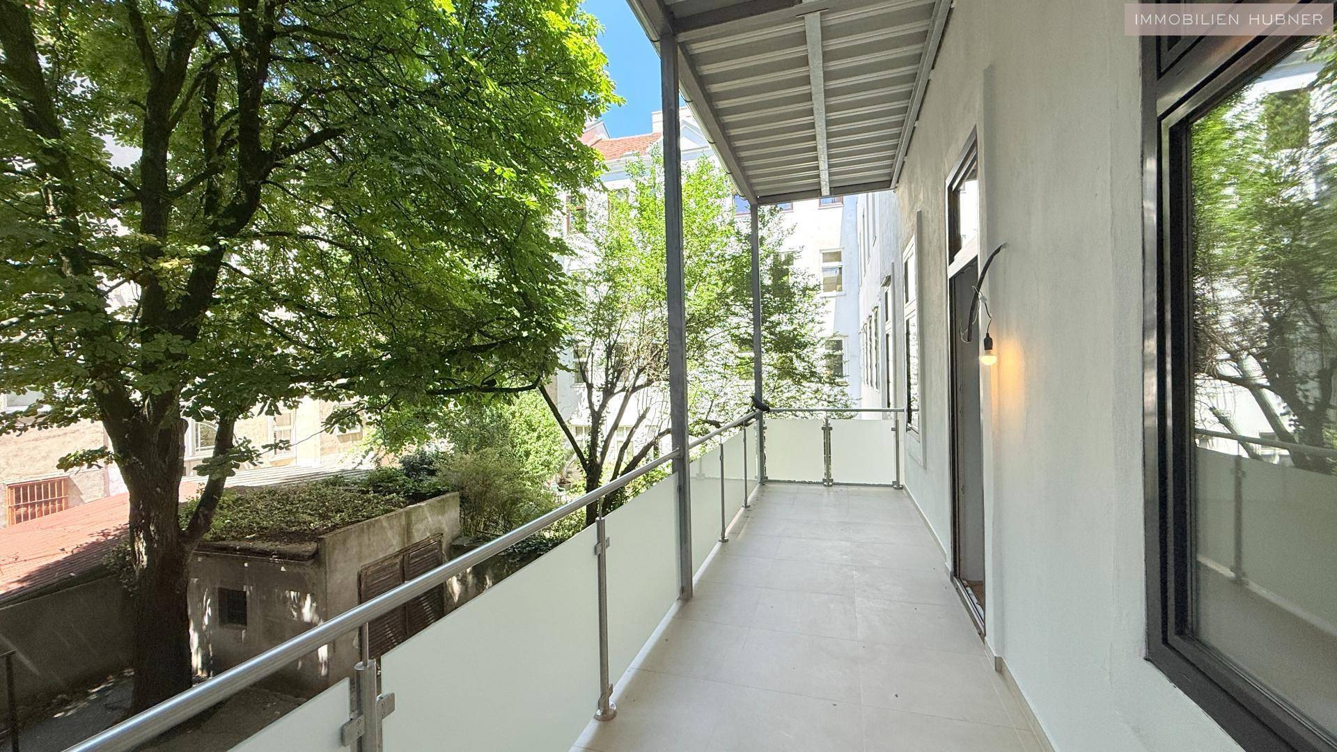 Langer Balkon mit Glasgeländer und Blick auf grüne Bäume in einem ruhigen Innenhof.