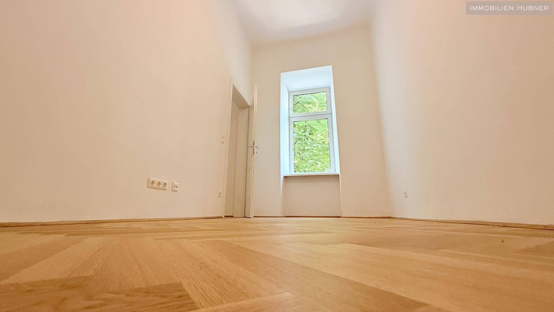 Helles Schlafzimmer mit Fischgrätparkettboden und einem großen Fenster, das viel Licht hereinlässt.