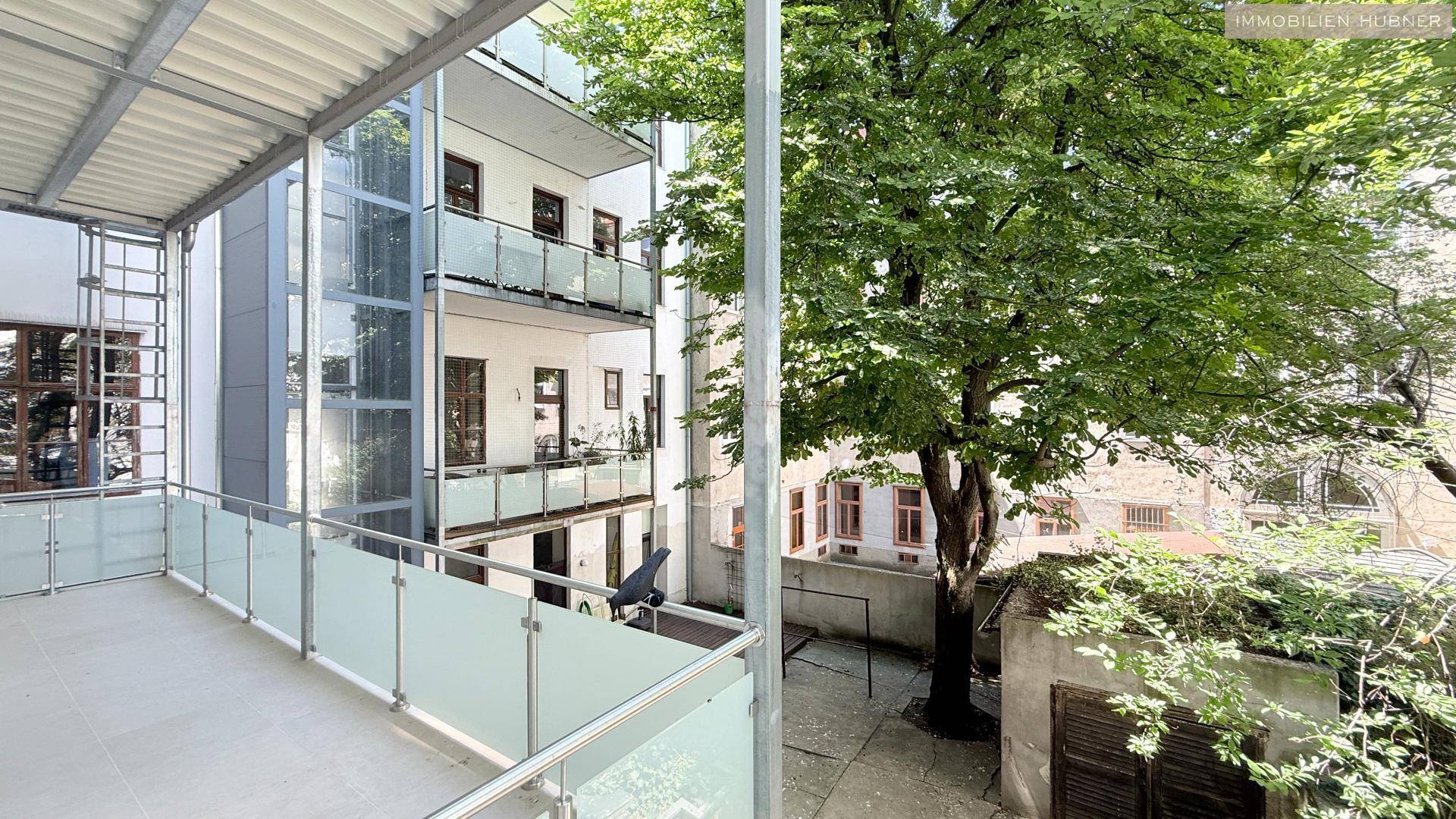 Balkon mit Blick auf weitere Balkone und einen begrünten Innenhof mit altem Baumbestand.