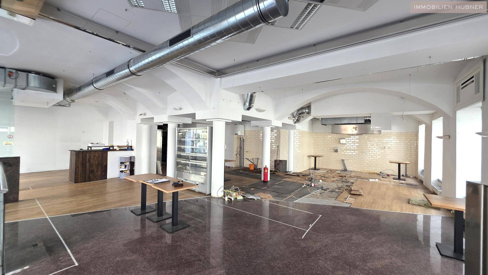 Großer Gewerberaum mit hohen Decken, sichtbaren Lüftungsrohren und unterschiedlichen Bodenbelägen.