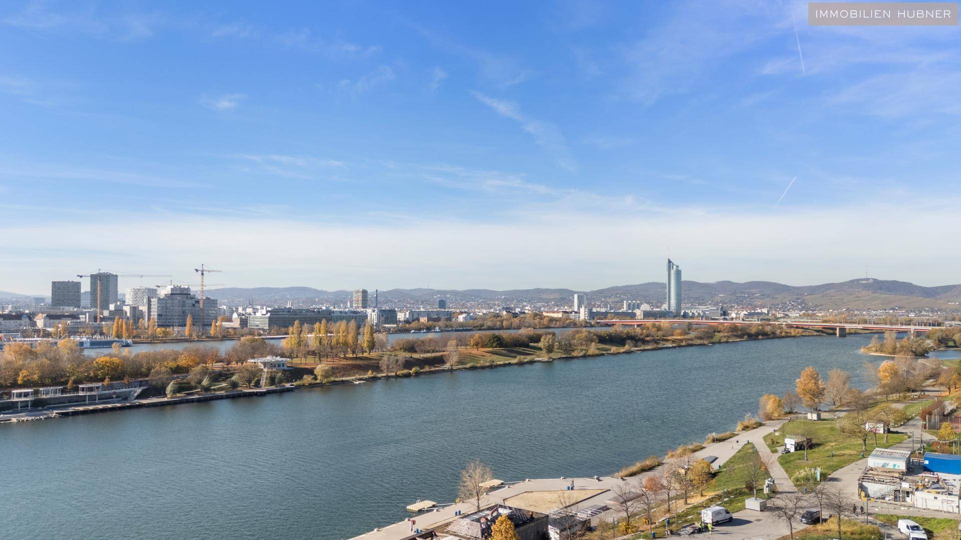 Panoramablick über die Stadt mit Fluss, modernen Gebäuden und Hügeln im Hintergrund unter blauem Himmel.
