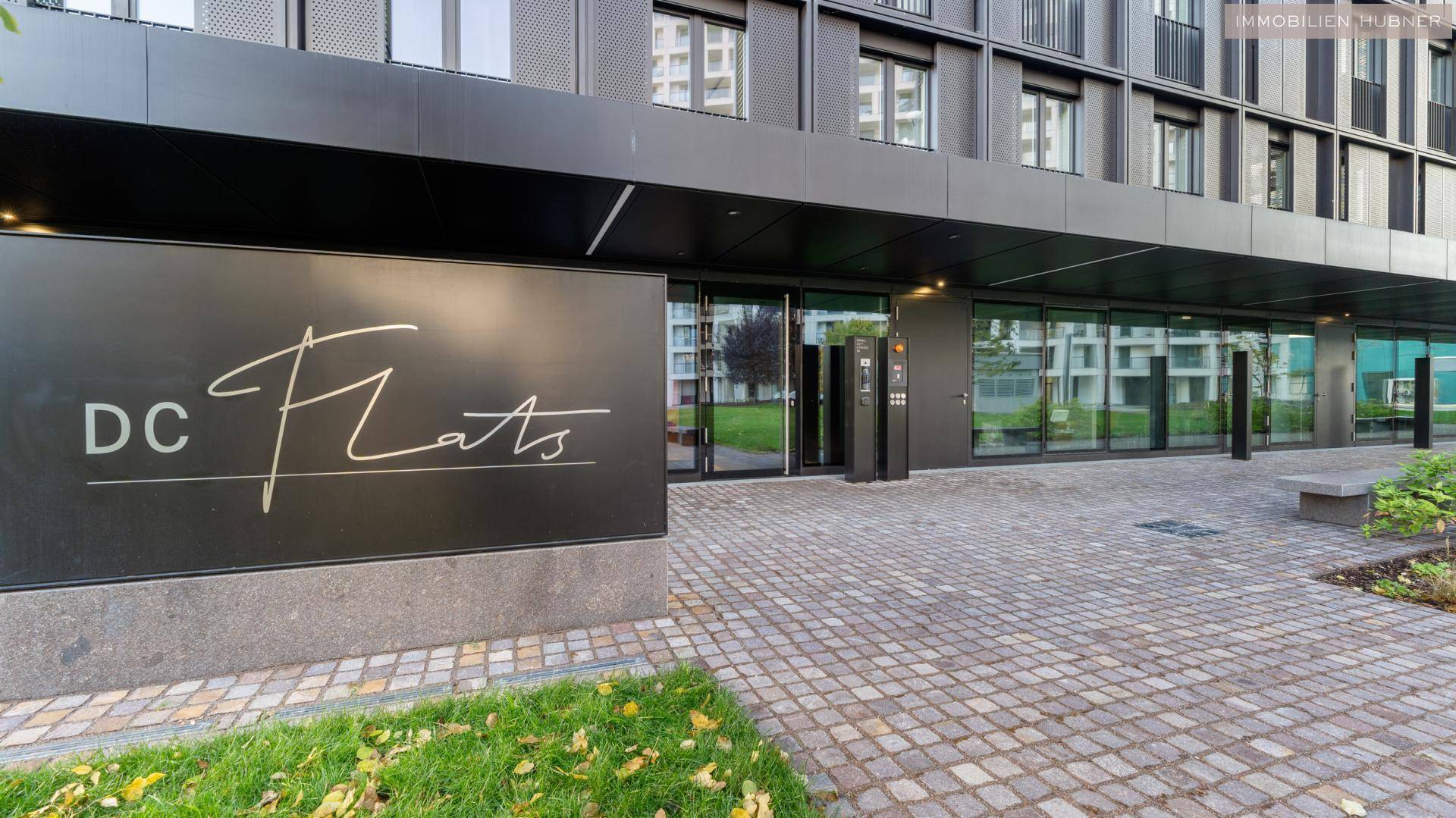 Eingangsbereich des modernen Gebäudes 'DC Flats' mit gepflastertem Vorplatz und Grünflächen.