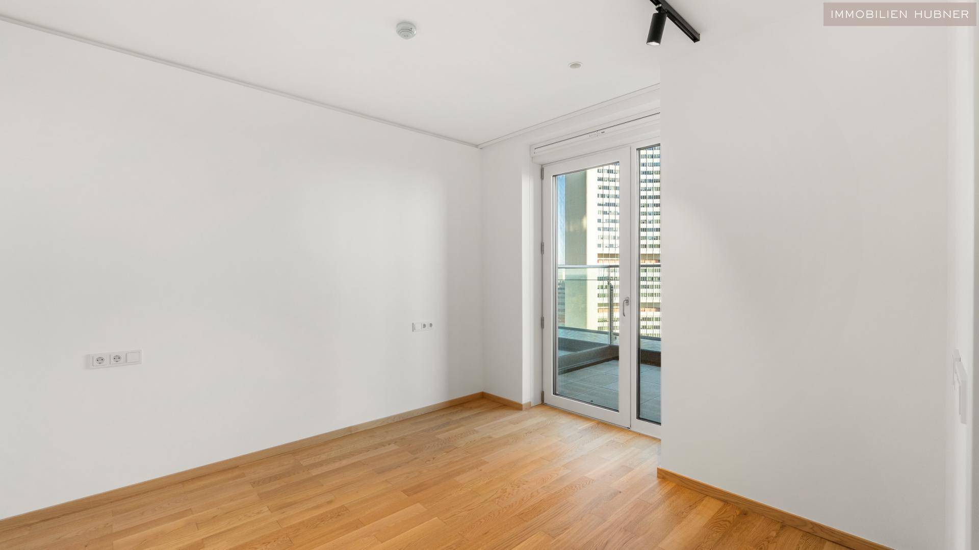 Leerer Raum mit Holzboden und großem Fenster, das Zugang zu einem Balkon bietet.