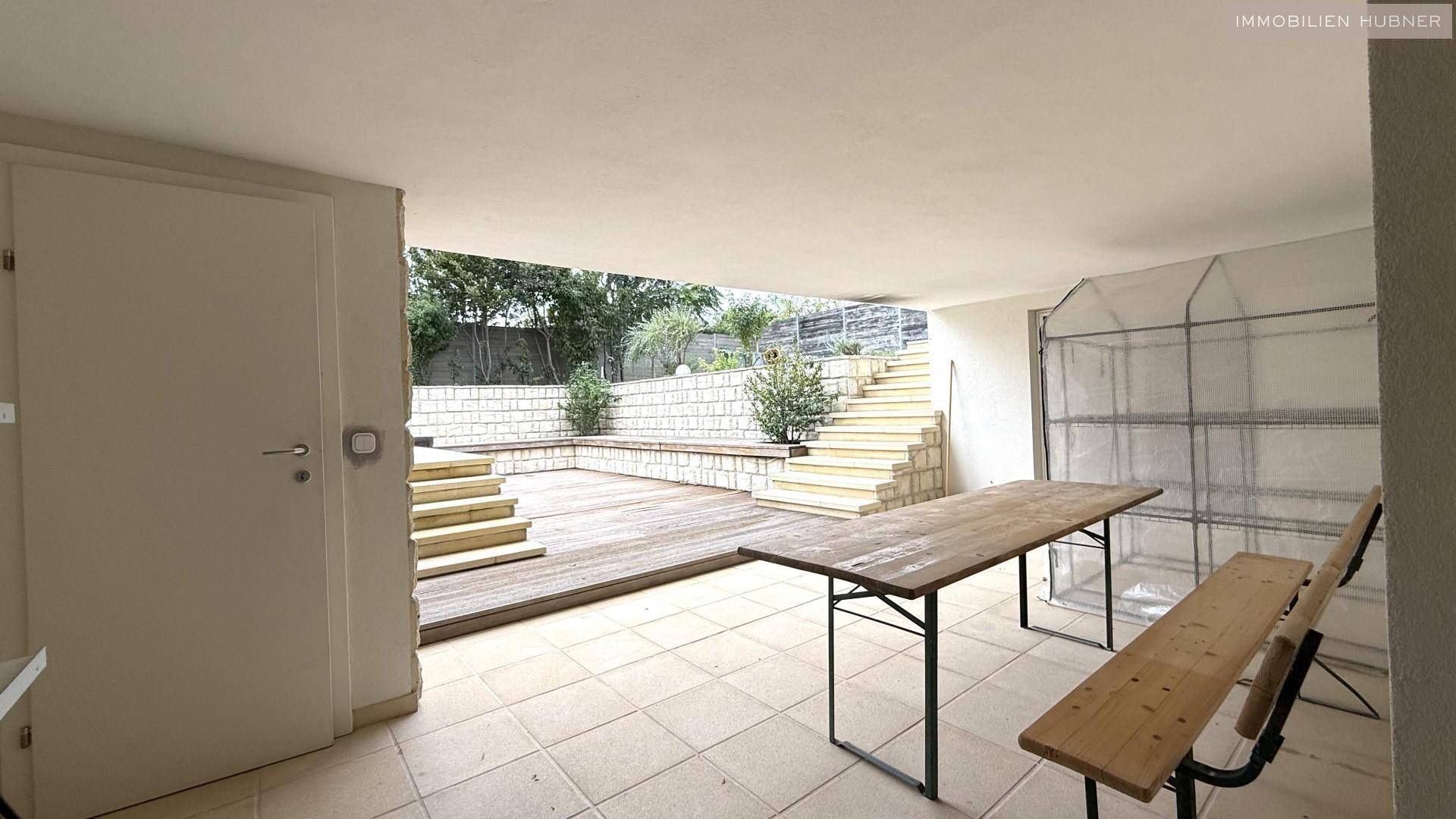 Überdachter Außenbereich mit Fliesenboden, Holztisch und Treppen zum Garten.