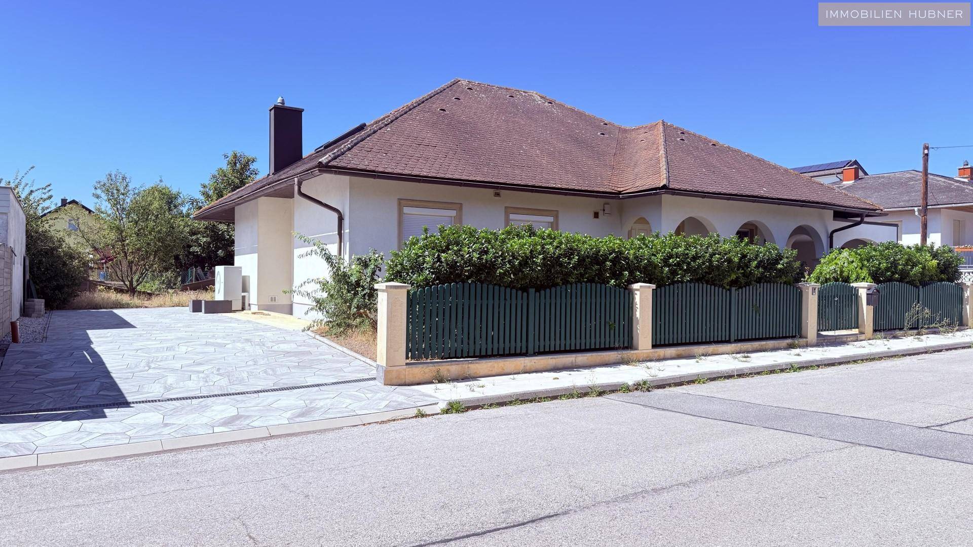 Seitenansicht des Hauses mit gepflasterter Auffahrt und grünem Zaun unter blauem Himmel.