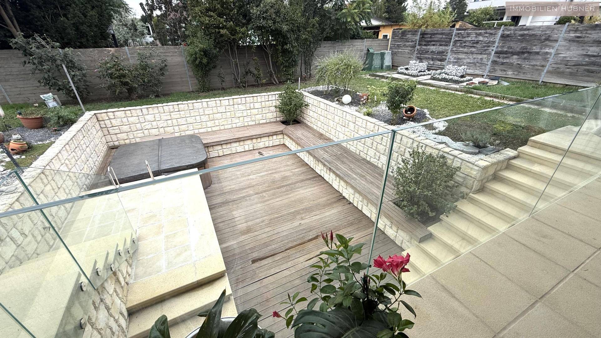 Versenkte Holzterrasse mit Whirlpool, Steinmauern und Glasgeländer im Gartenbereich.