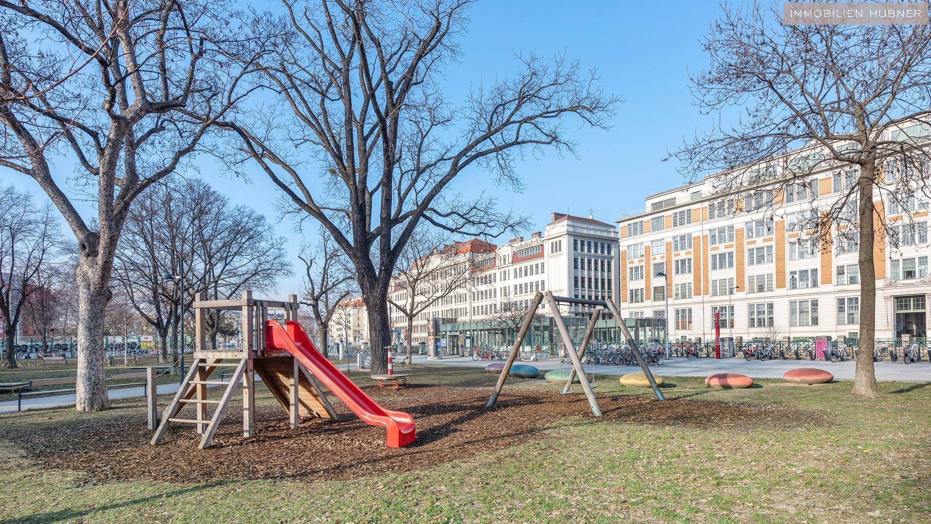 Öffentlicher Spielplatz mit Rutsche und Schaukeln in einem Stadtpark mit Bäumen.