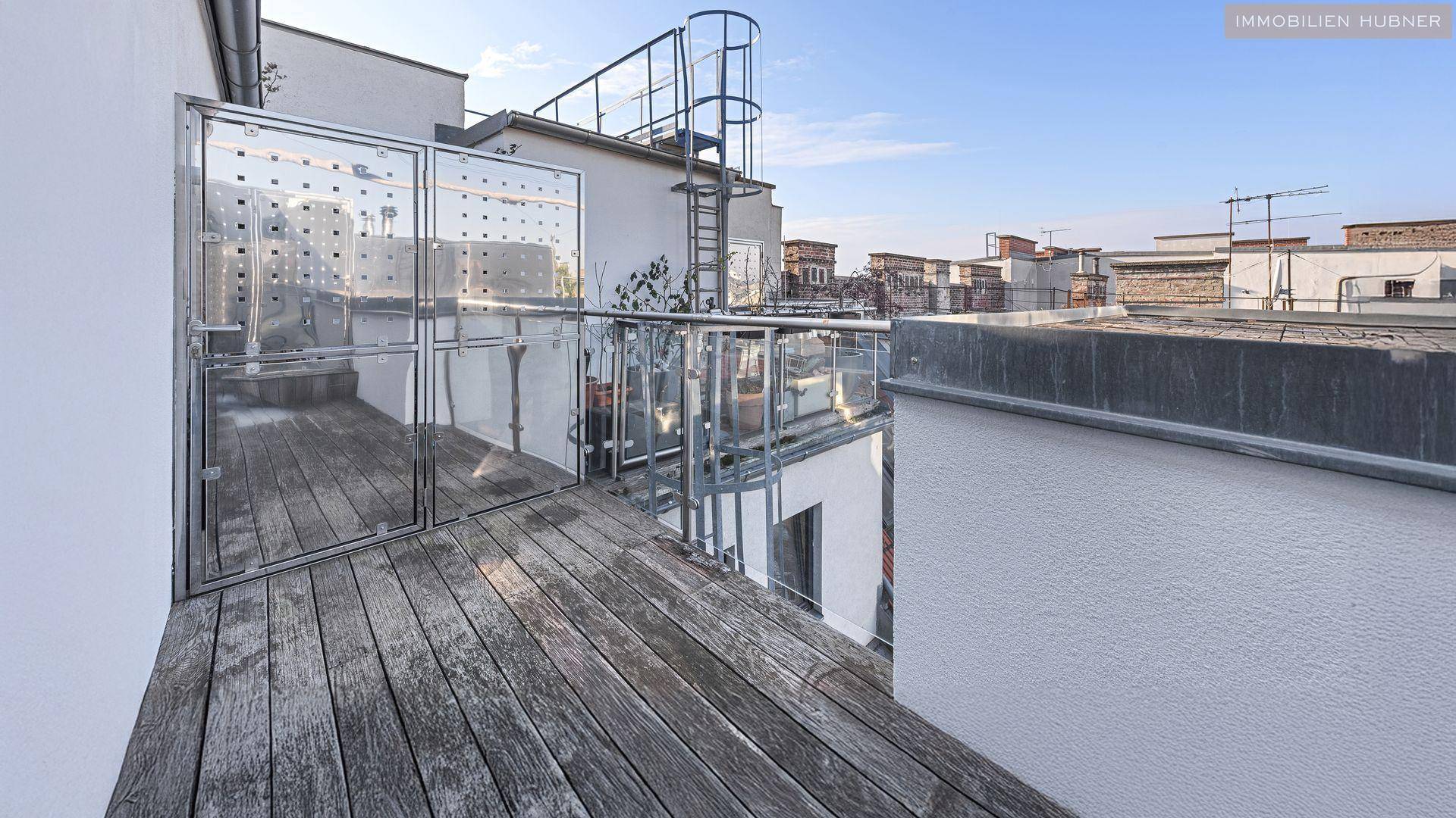 Geräumige Dachterrasse mit Holzboden, Glasgeländer und Blick über die Dächer der Stadt.