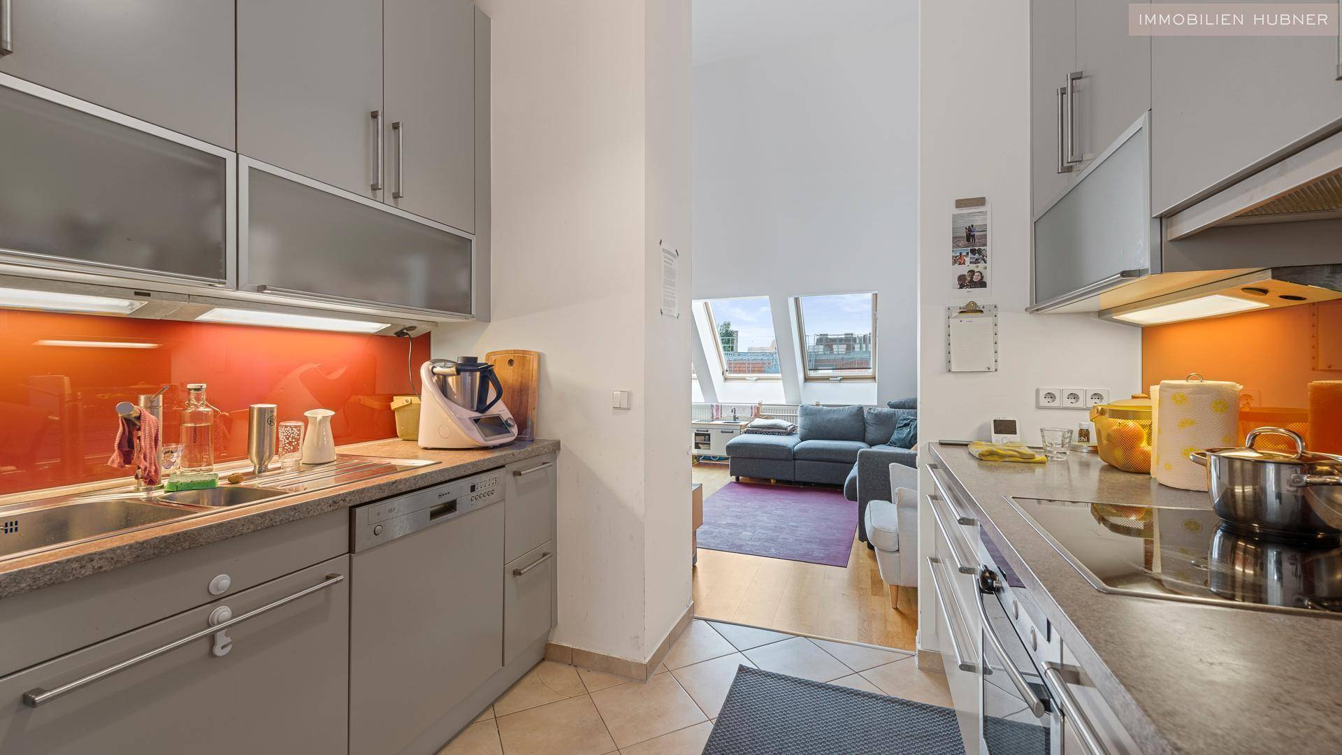 Moderne Küche mit grauen Schränken, orangefarbener Rückwand und Blick auf den Wohnbereich mit Dachfenstern.