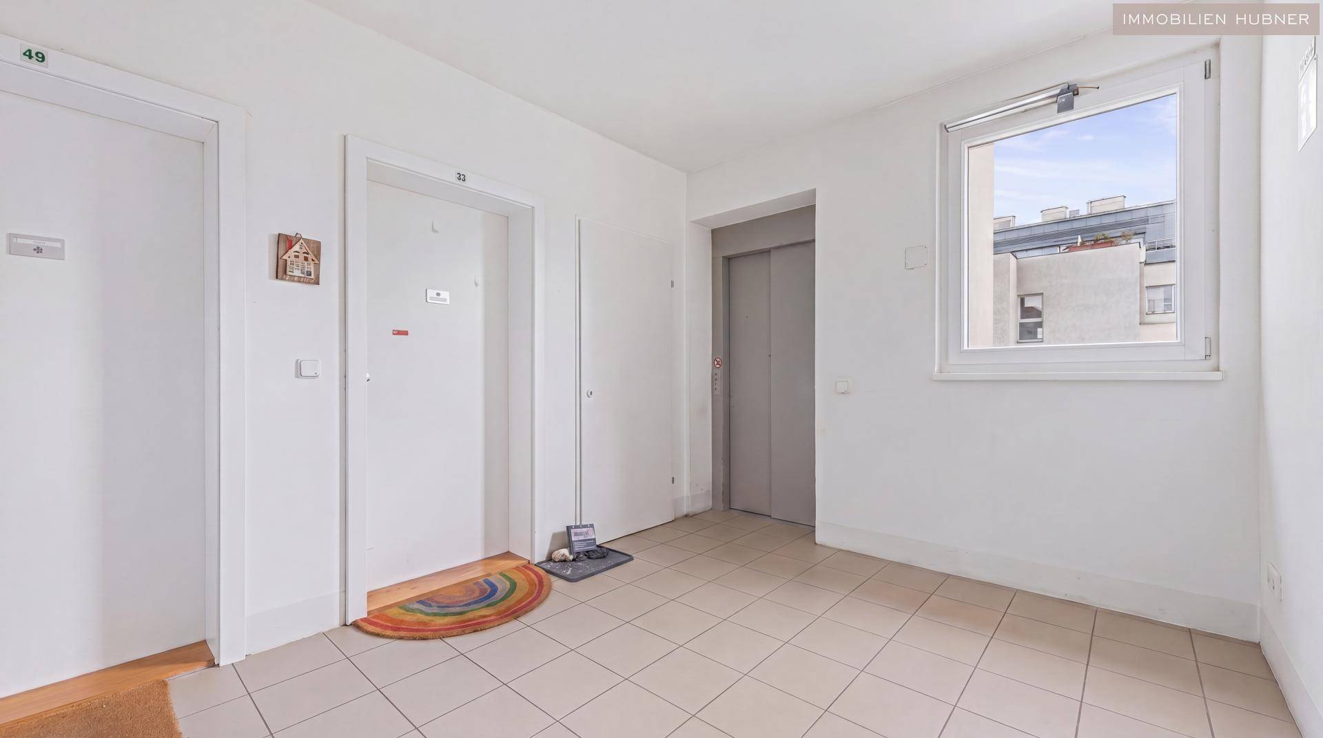 Heller Hausflur mit Fliesenboden, zwei Wohnungstüren, einem Aufzug und einem Fenster.