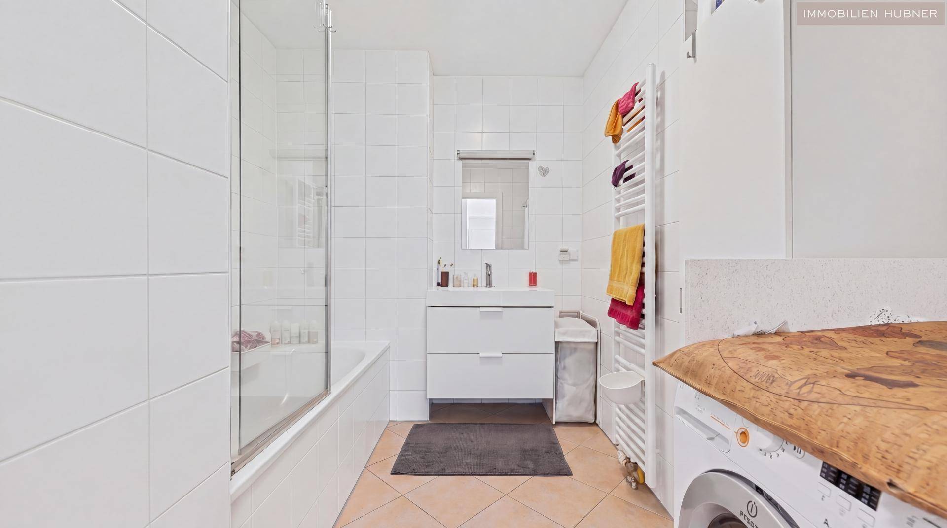 Modernes Badezimmer mit weißen Fliesen, Badewanne mit Duschwand und Waschmaschine, gut beleuchtet.