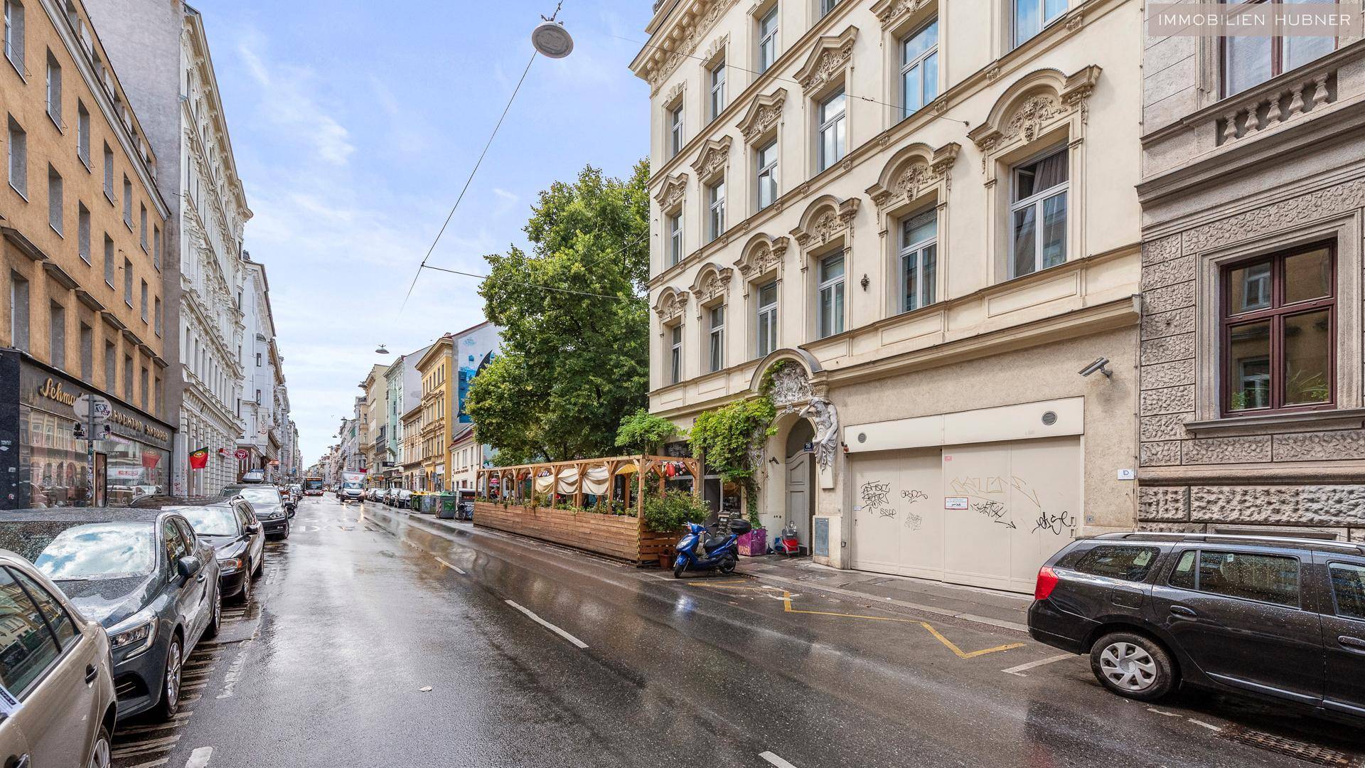 Belebtes Stadtbild mit geparkten Autos, historischen Gebäuden und einer Außenterrasse eines Restaurants.