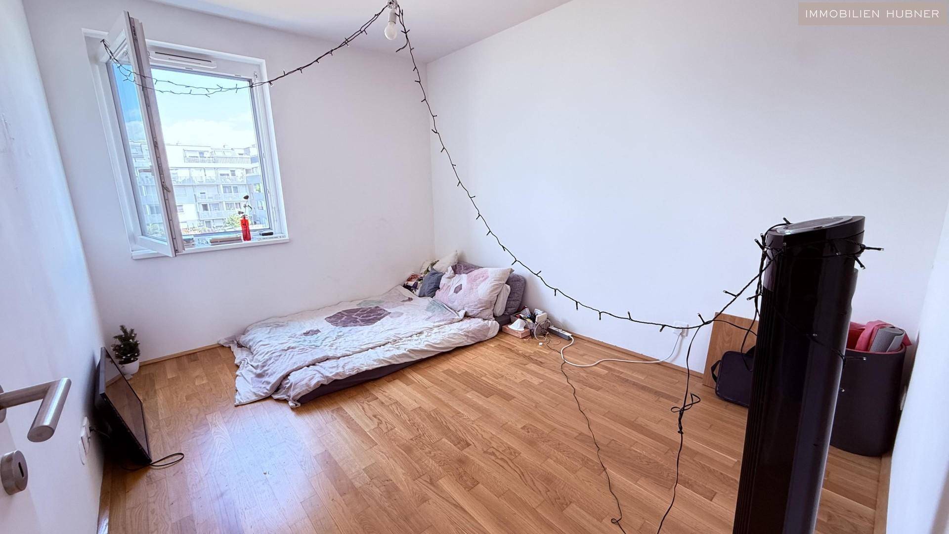 Schlafzimmer mit Holzboden, großem Fenster und einfacher Einrichtung.
